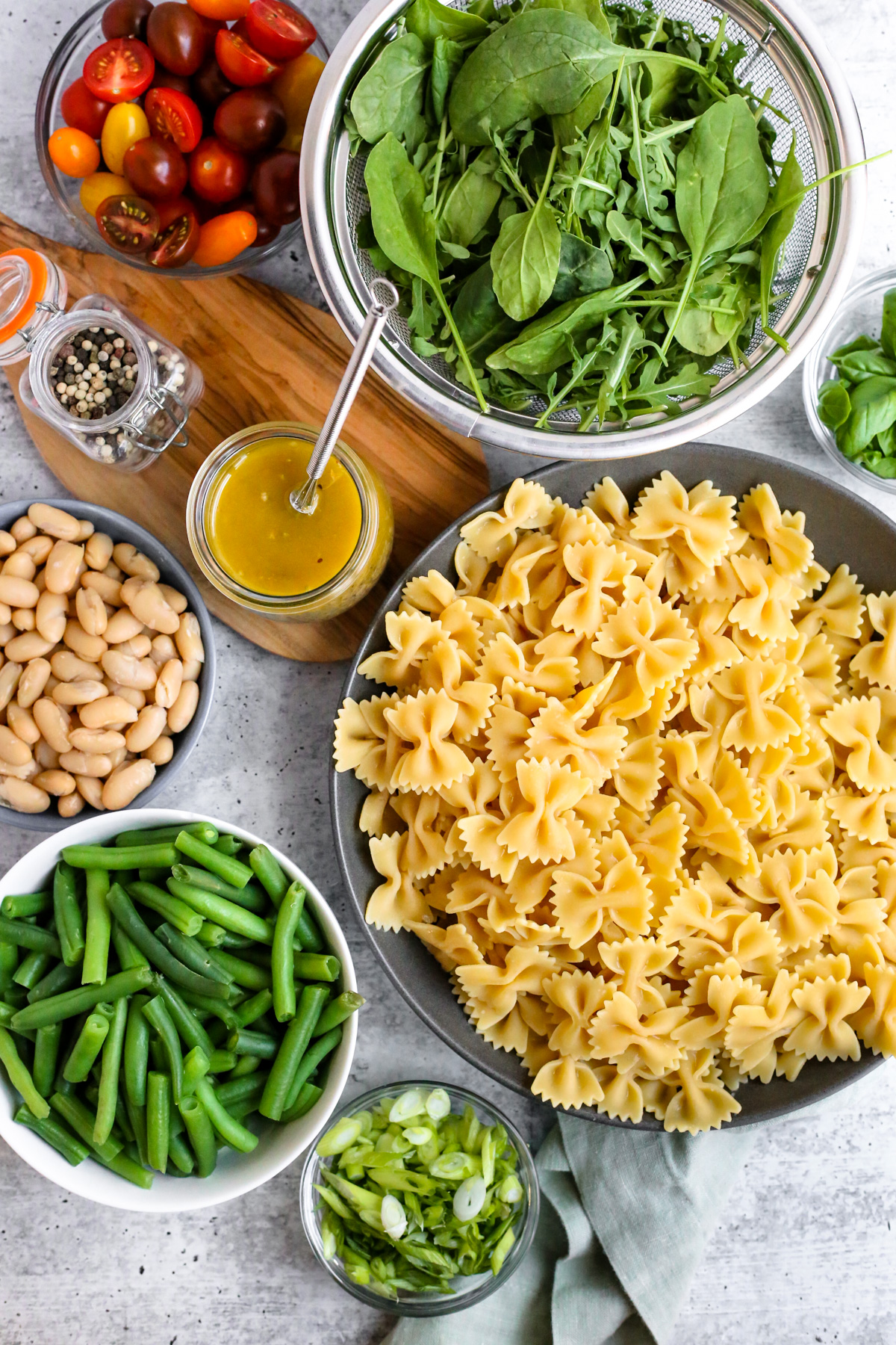 Flatlay style image of the ingredients needed for a beans and greens pasta salad, including cooked bowtie pasta, sliced green onions and cherry tomatoes, blanched and trimmed green beans, canned cannellini beans, fresh salad greens and basil, and a homemade red wine vinaigrette dressing