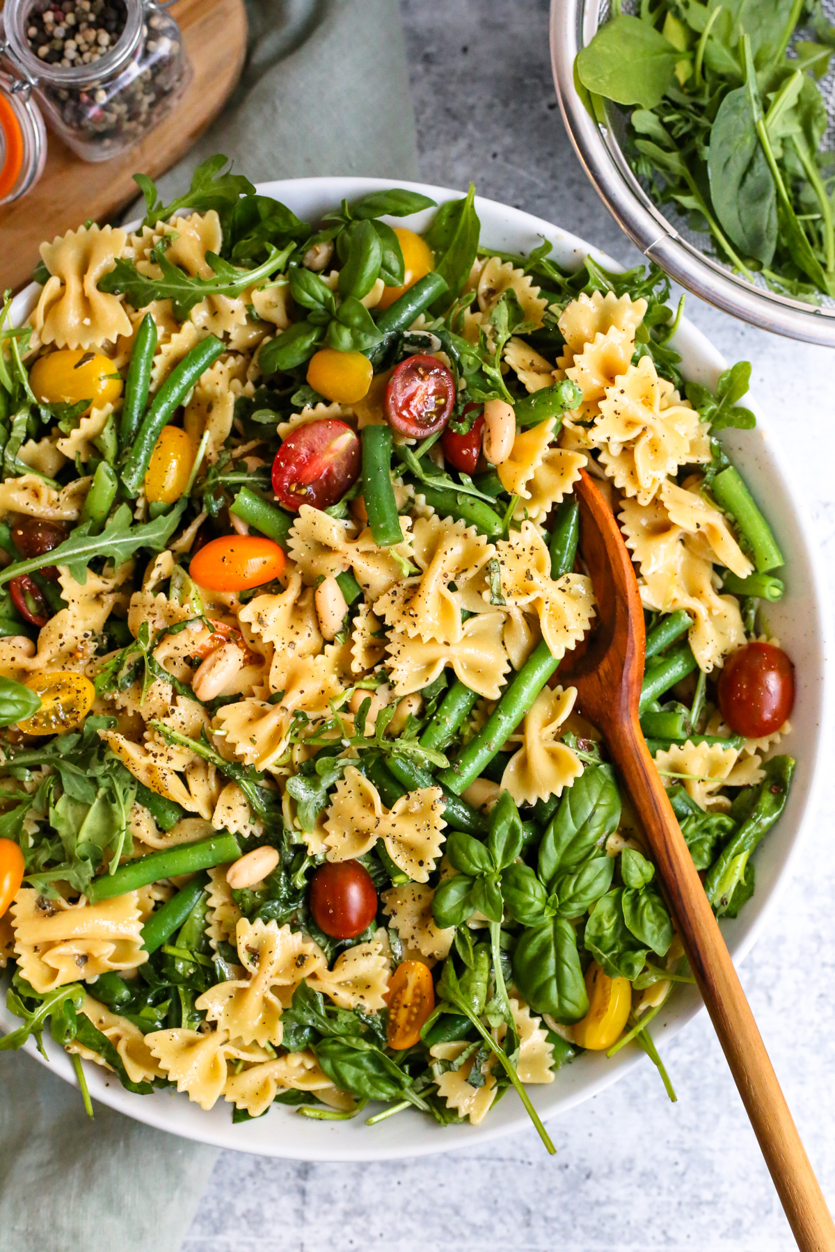 An overhead view of a large white serving bowl filled with a colorful pasta salad using bowtie pasta, fresh salad greens, green beans, cherry tomatoes, white beans, and fresh basil, coated lightly in a homemade vinaigrette salad dressing and garnished with cracked black pepper