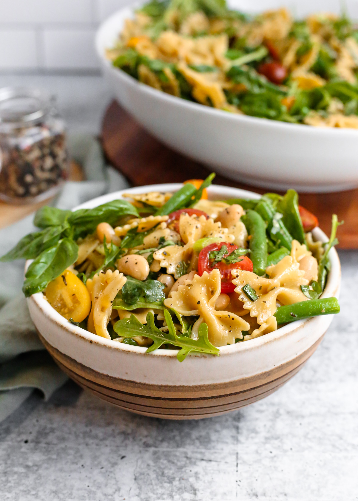 Angled view of a bowl of beans and greens pasta salad, using bowtie pasta, cherry tomatoes, green beans, white beans, sliced green onions, and a light vinaigrette dressing. Behind the bowl is a larger serving bowl on a wooden platter