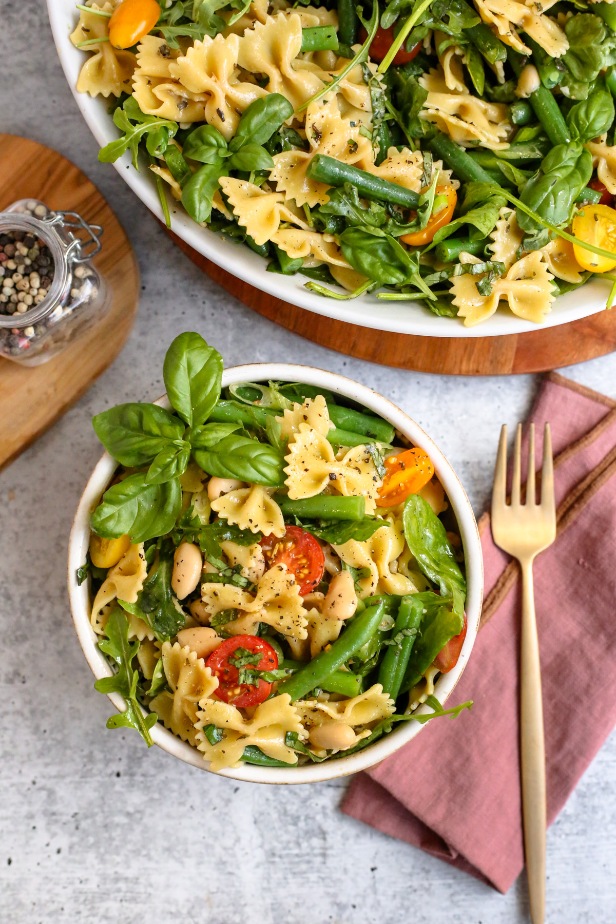 Overhead view of a bowl of fresh pasta salad made with beans, greens, cherry tomatoes, fresh basil, and blanched green beans. The serving is garnished with extra basil and black pepper and it's placed on a kitchen countertop next to a fork and napkin, appearing ready to eat