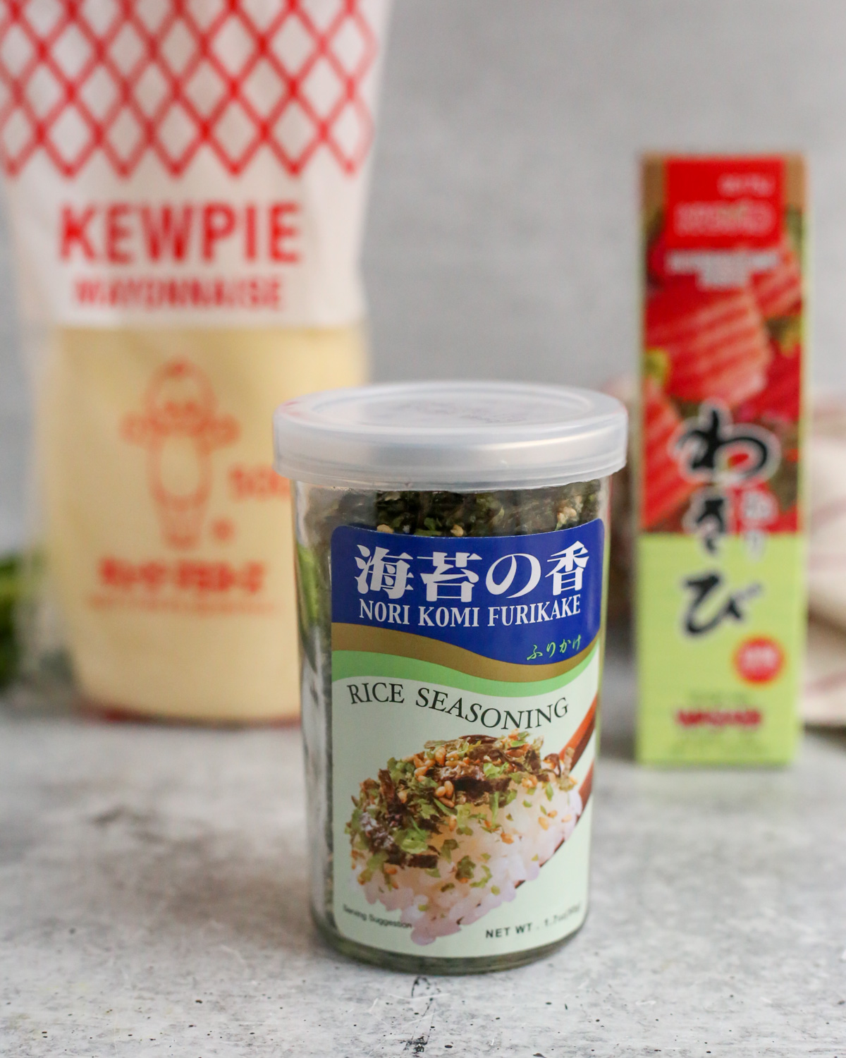 A close up view of a bottle of nori kumi Furikake, with the label for "rice seasoning", displayed on a kitchen countertop with an unopened bottle of kewpie mayo and unopened tube of wasabi paste visible in the background
