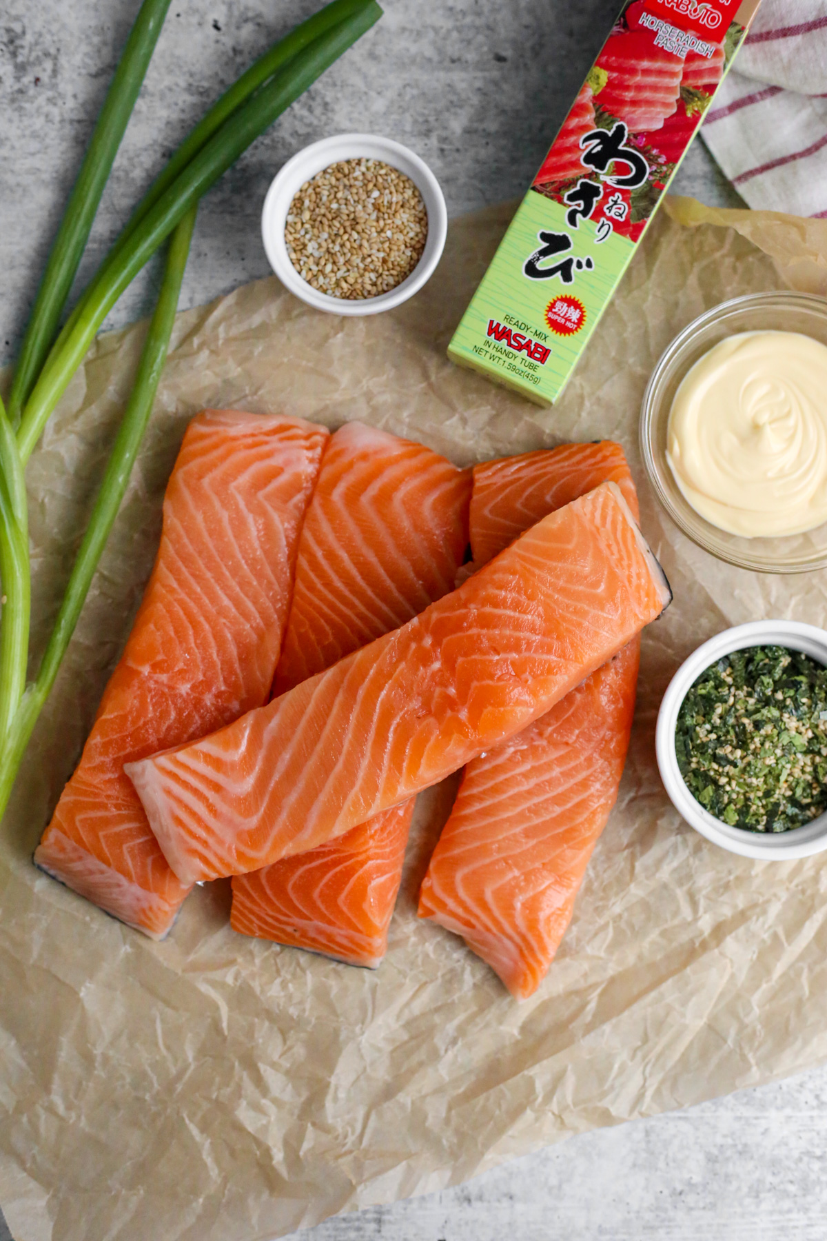Flatlay style image with the ingredients for this salmon recipe, including four sliced, skin-on salmon fillets, green onions, a ramekin with sesame seeds, a ramekin with Furikake, an unopened tube of wasabi paste, and small glass dish with mayonnaise 