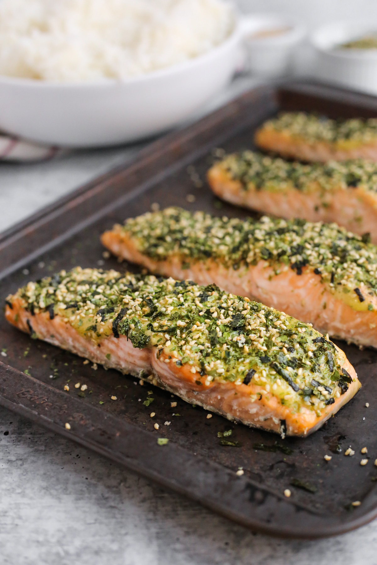 A sheet pan with patina displays four cooked pieces of Furikake salmon on a kitchen countertop, with a bowl of steamed white rice cooked and ready to serve in the background