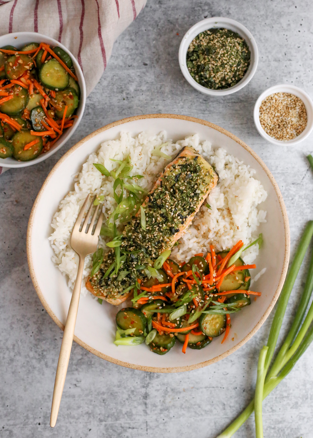 Overhead view of a bowl of steamed white rice, topped with a fillet of salmon coated in Furikake seasoning, garnished with sliced green onions and a side of sliced vegetables, ready to be serve with a brass colored fork resting on one side of the dish