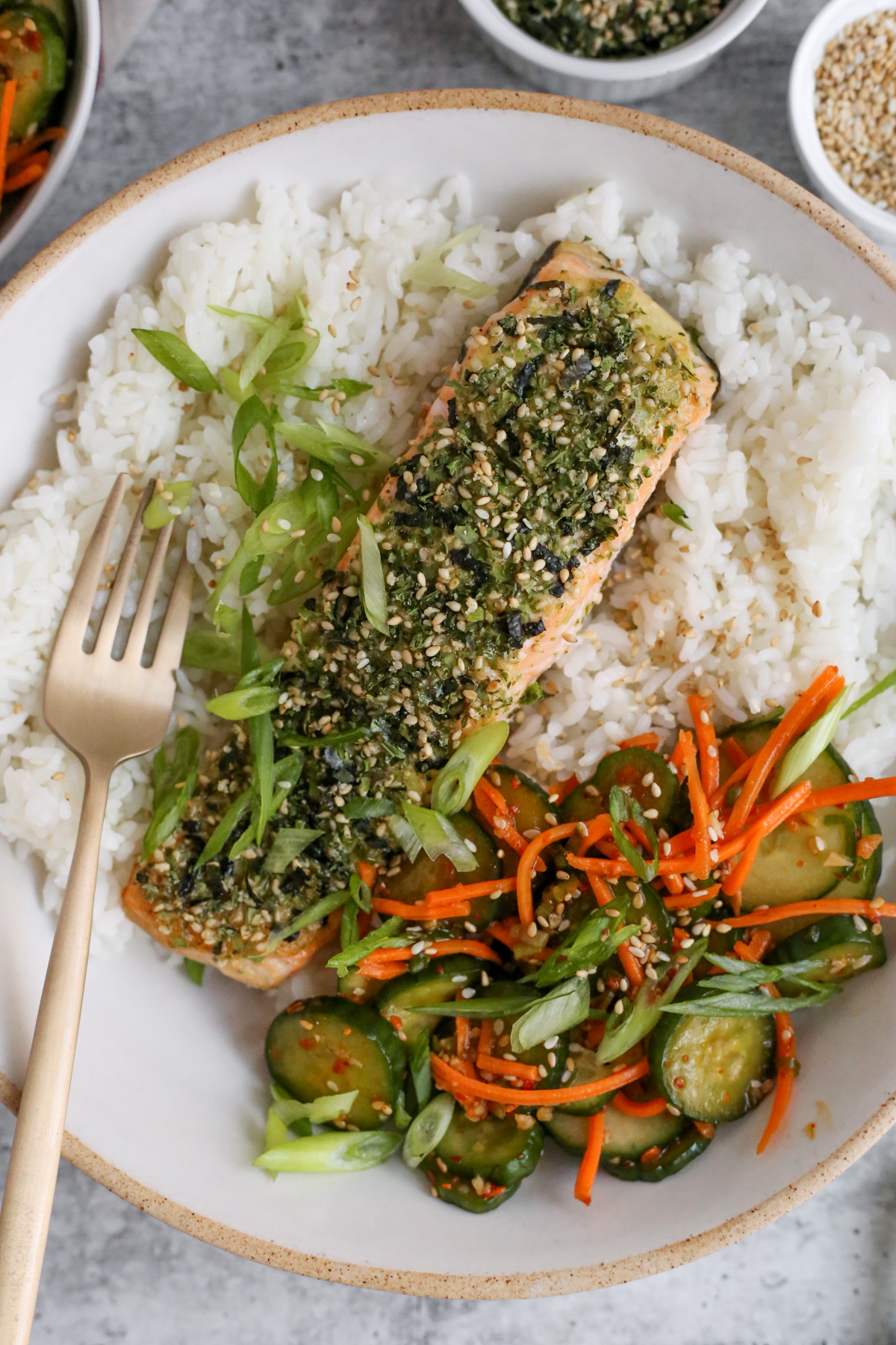 Overhead view of a bowl of white rice topped with a fillet of Furikake salmon, served with green onions and sliced cucumbers on the side