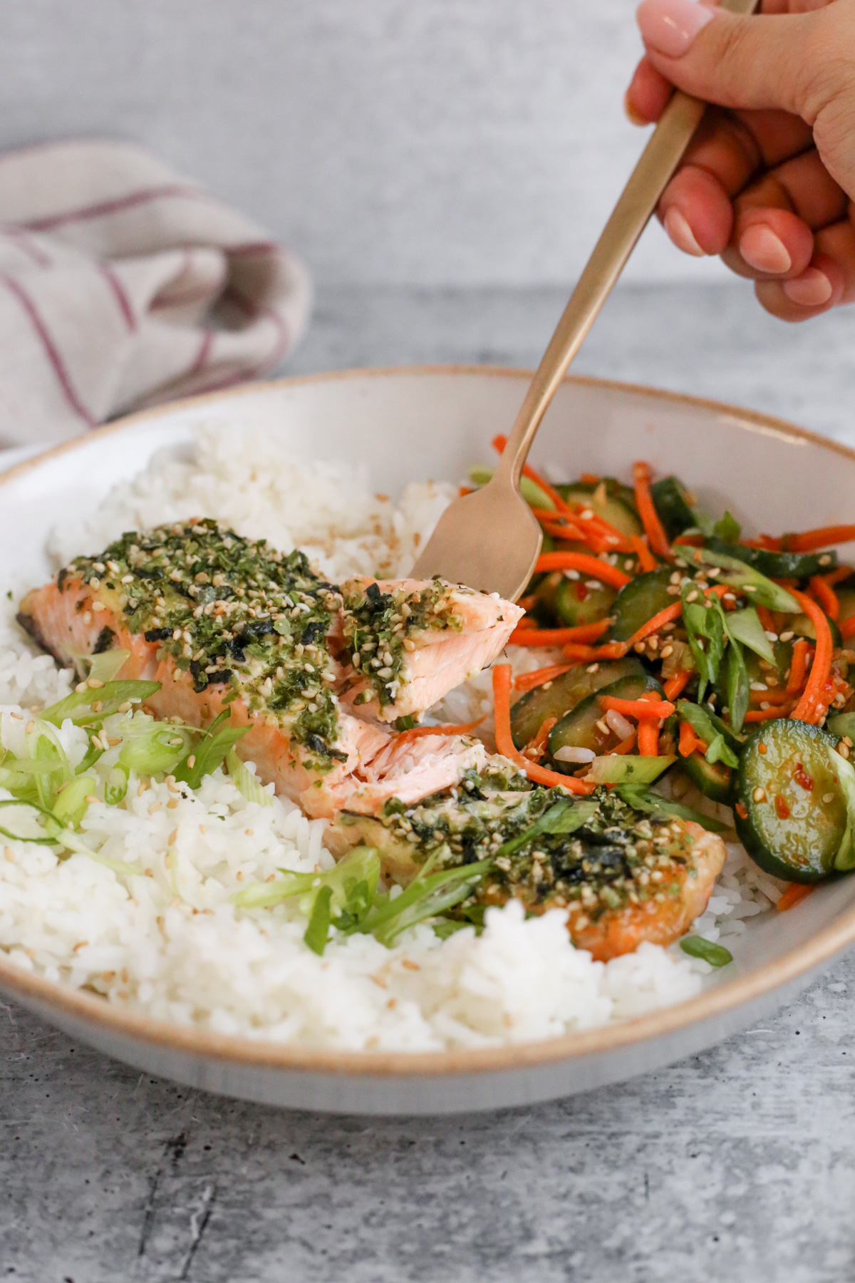 A woman's hand holds a brass colored fork and reaches into a bowl of rice and Furikake salmon, pulling again a bite of salmon that looks flaky, with various sliced vegetables as sides