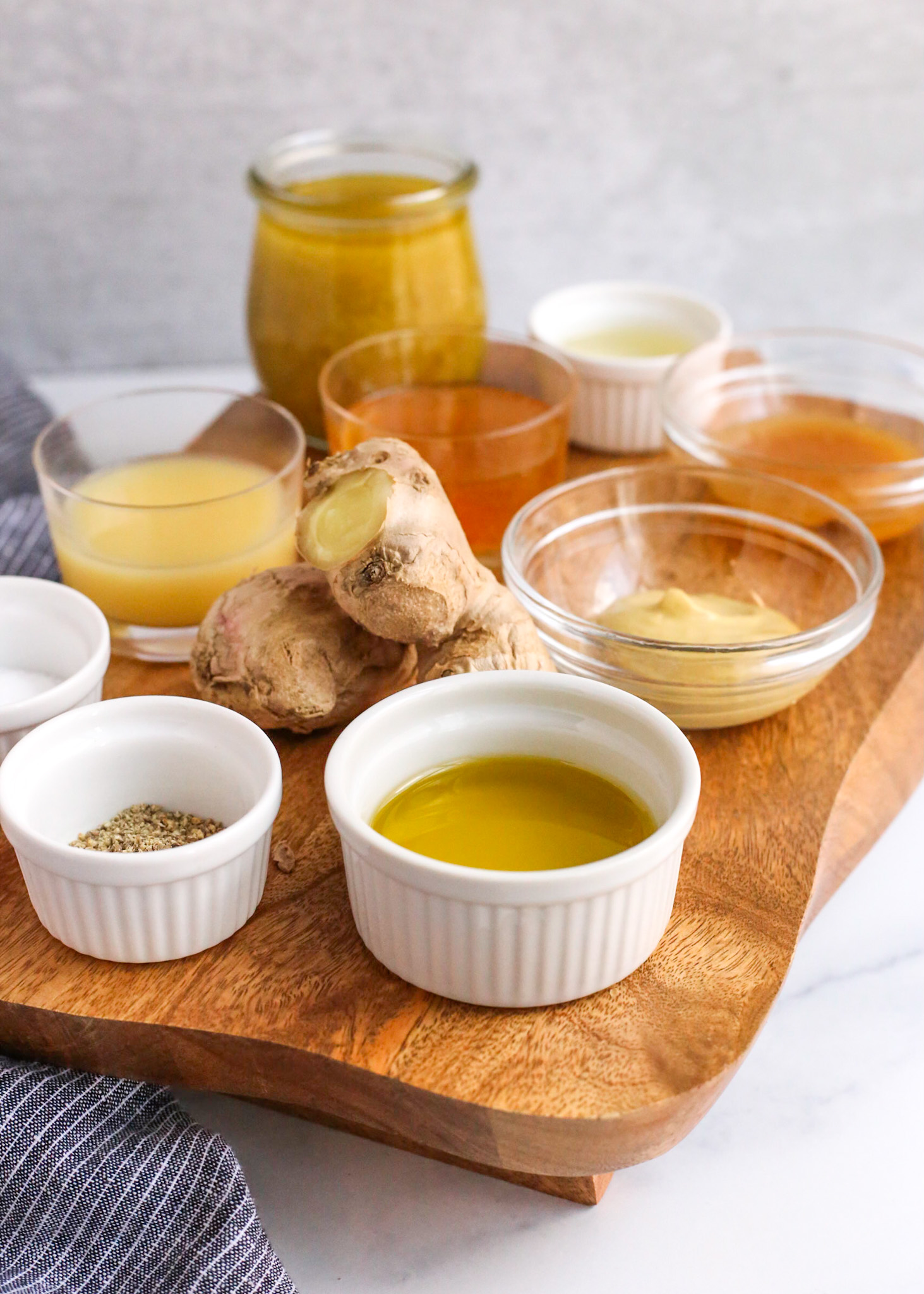 Angled image of a kitchen countertop with a wooden serving board displaying the ingredients for a yuzu vinaigrette salad dressing in small white or glass ramekins, with a knob of fresh ginger in the center of the board