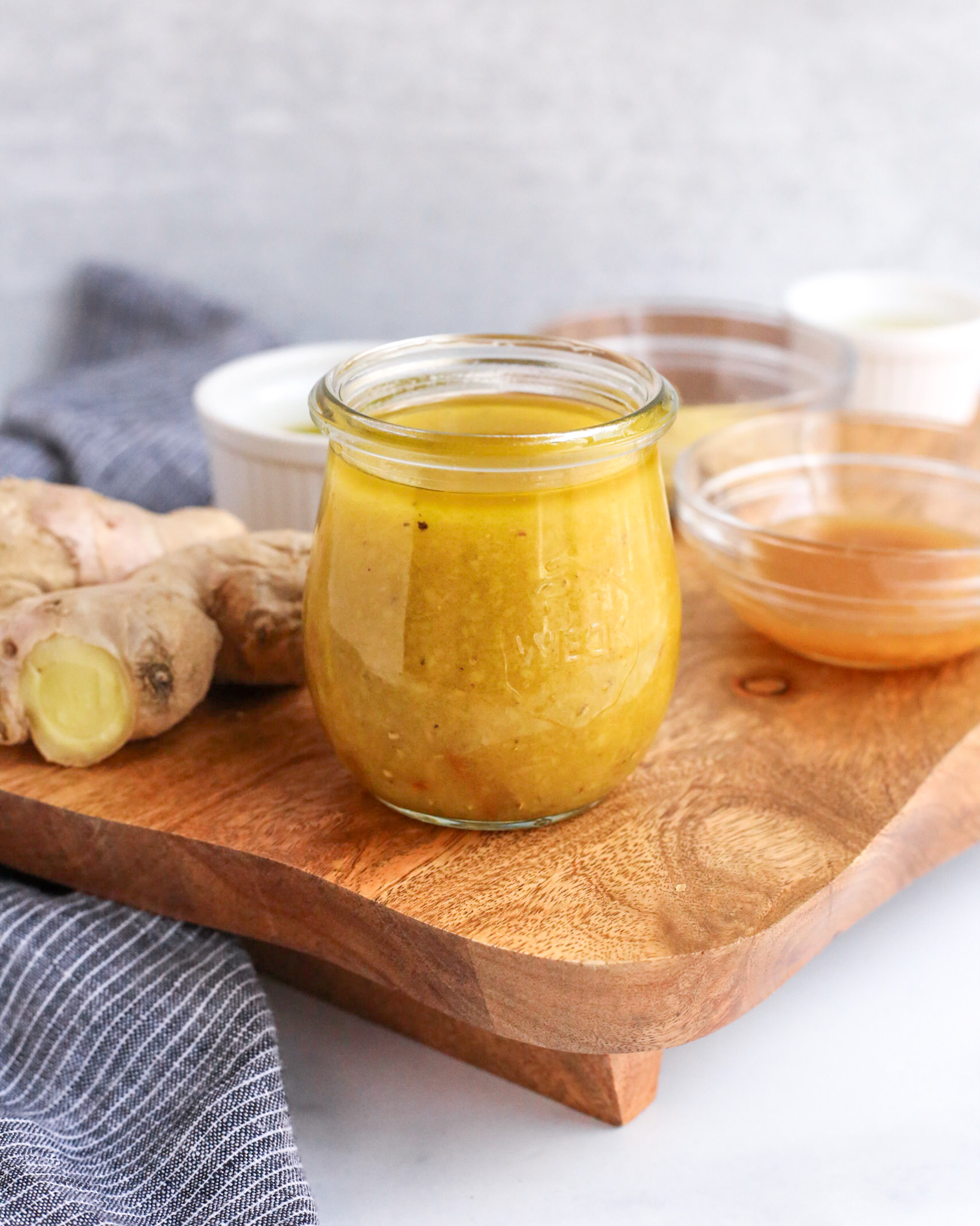 A closeup side view of a small glass jar filled to the top with a yuzu vinaigrette that is golden yellow in color with pieces of pepper, fresh ginger, and yuzu rind suspended in it