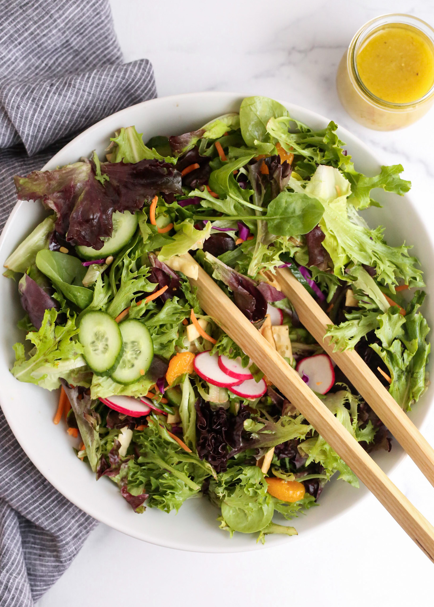 Overhead view of a mixed greens salad in a large white serving bowl with wooden salad tongs set on the side of the bowl. A blue striped linen and small jar of yuzu salad dressing are included nearby