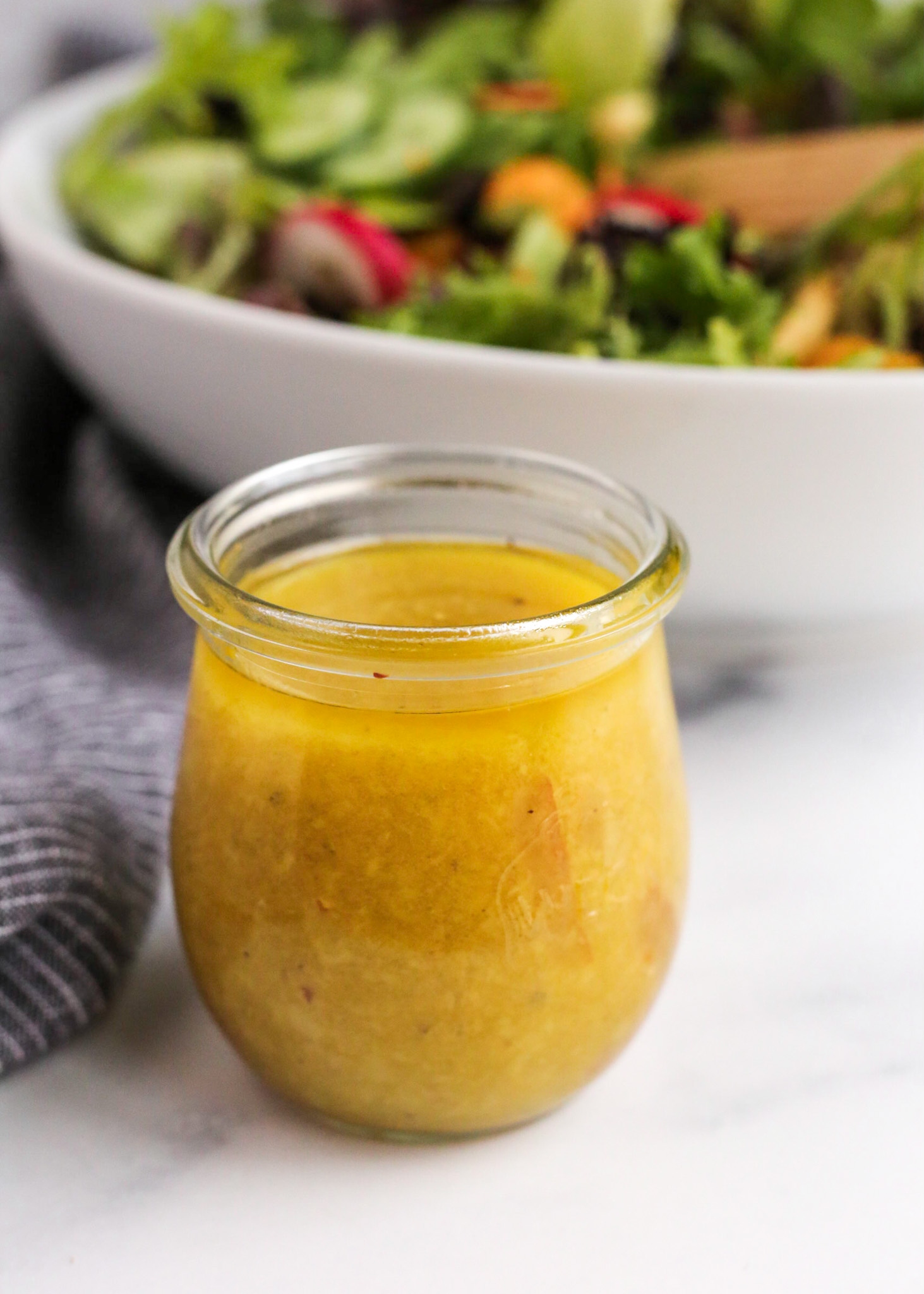 A close view of a small glass jar with a yuzu salad dressing inside, displayed on a kitchen countertop with a mixed greens salad visible in the background