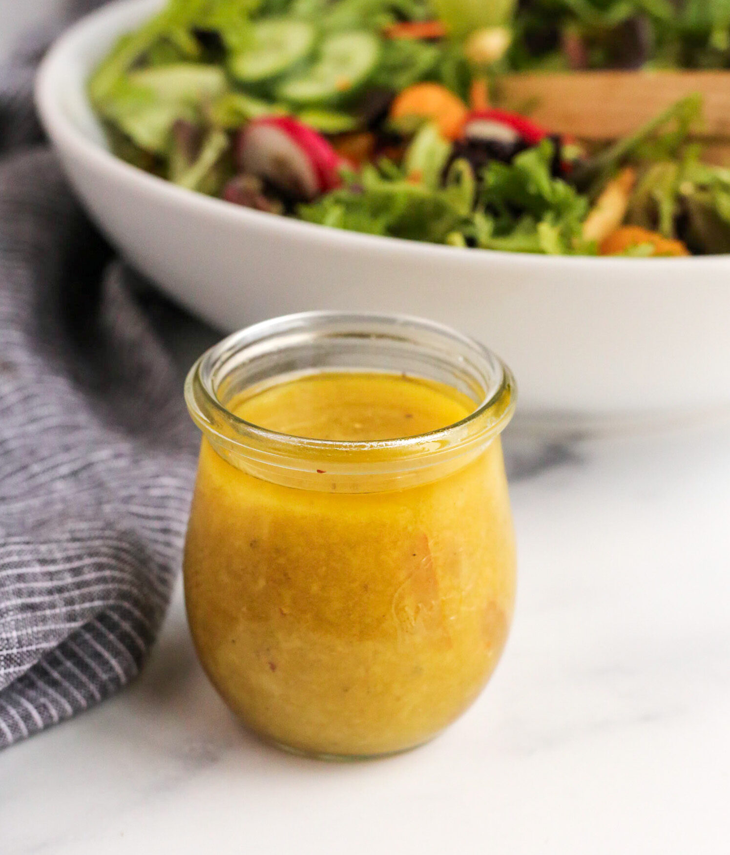 A small, clear glass jar without a lid sits on a kitchen countertop, filled to the top with a bright citrusy yuzu salad dressing with the mixed greens salad in the background