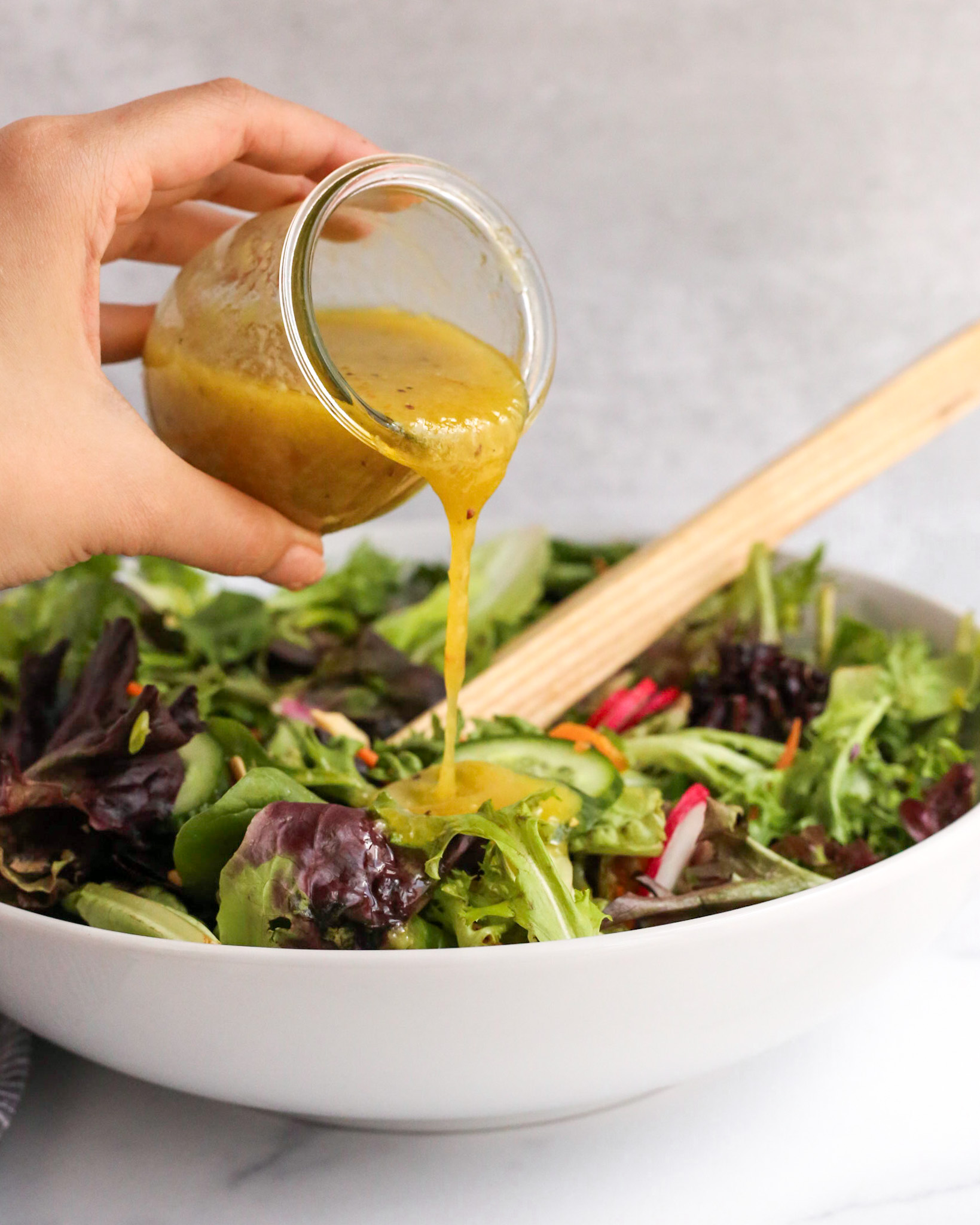 A woman's hand holds a small clear glass jar of yuzu dressing and pours it over a mixed greens salad