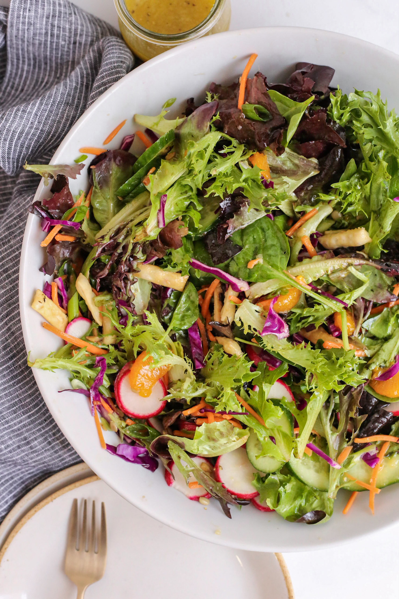 Overhead view of a large serving bowl full of a mixed greens salad with shredded carrots and cabbage, sliced green onions and radishes, sunflower seeds, wonton strips, and a yuzu dressing on a kitchen countertop