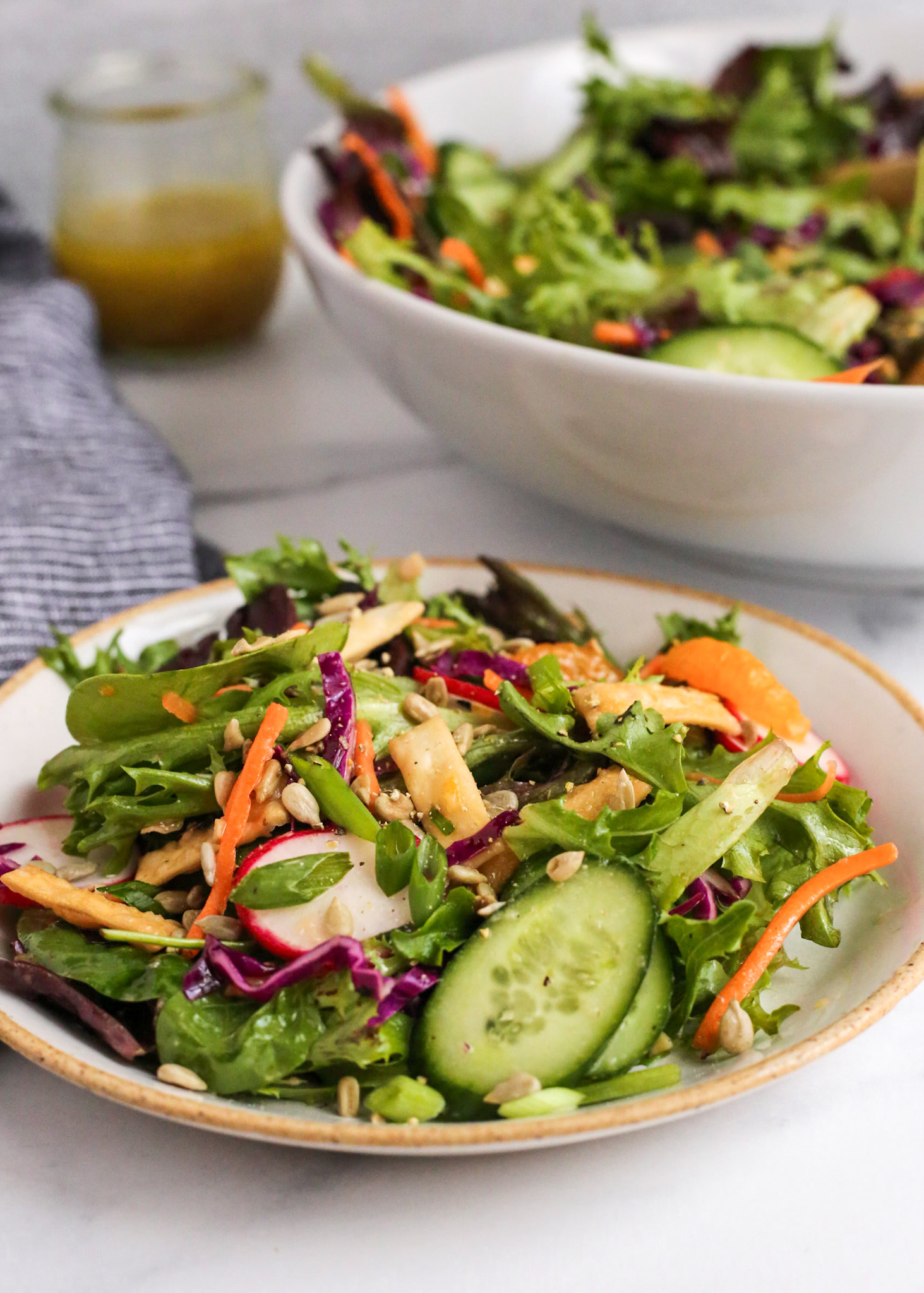 Angled view of a side salad with leafy greens, mandarin oranges, and colorful sliced vegetables, served on a beige ceramic salad plate with extra yuzu dressing and the serving bowl of salad in the background