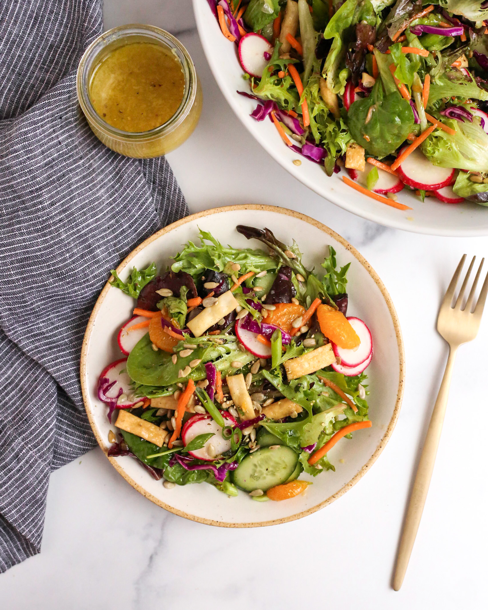 Overhead view of a plated mixed greens salad with yuzu salad dressing, served on a beige ceramic salad plate with a brass fork nearby, along with the serving bowl full of salad and extra dressing nearby