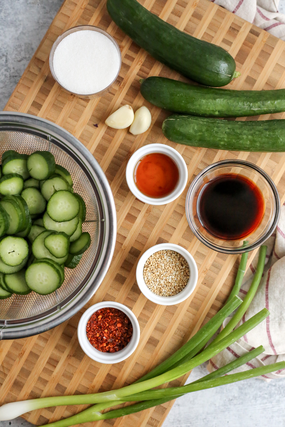 A wooden butcher block cutting board displays a stainless steel colander filled with sliced cucumbers with the sauce ingredients for oi muchim, spicy Korean cucumber salad, in small ramekins and dishes, with extra Persian cucumbers, garlic cloves, and green onions arranged within the frame