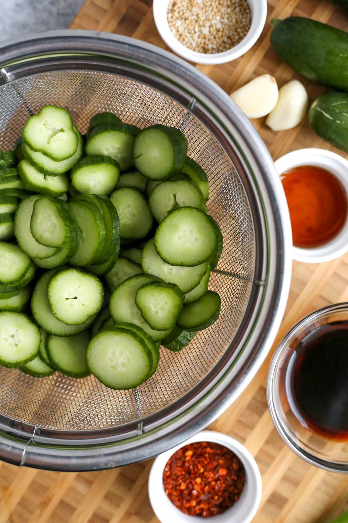 Overhead view of a stainless steel colander filled with thinly sliced cucumber and the prepared ingredients to make oi muchim, or spicy korean cucumber side