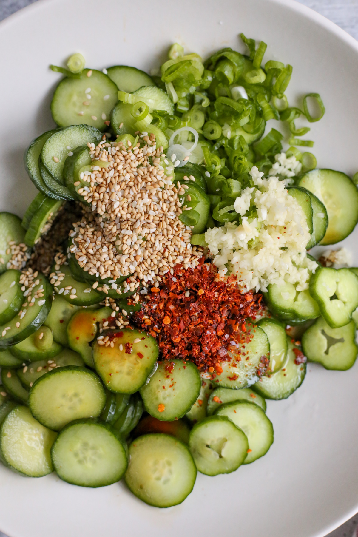 Overhead view of a mixing bowl filled with sliced cucumbers, minced garlic, toasted sesame seeds, red pepper flakes (gochugaru), and sliced green onion