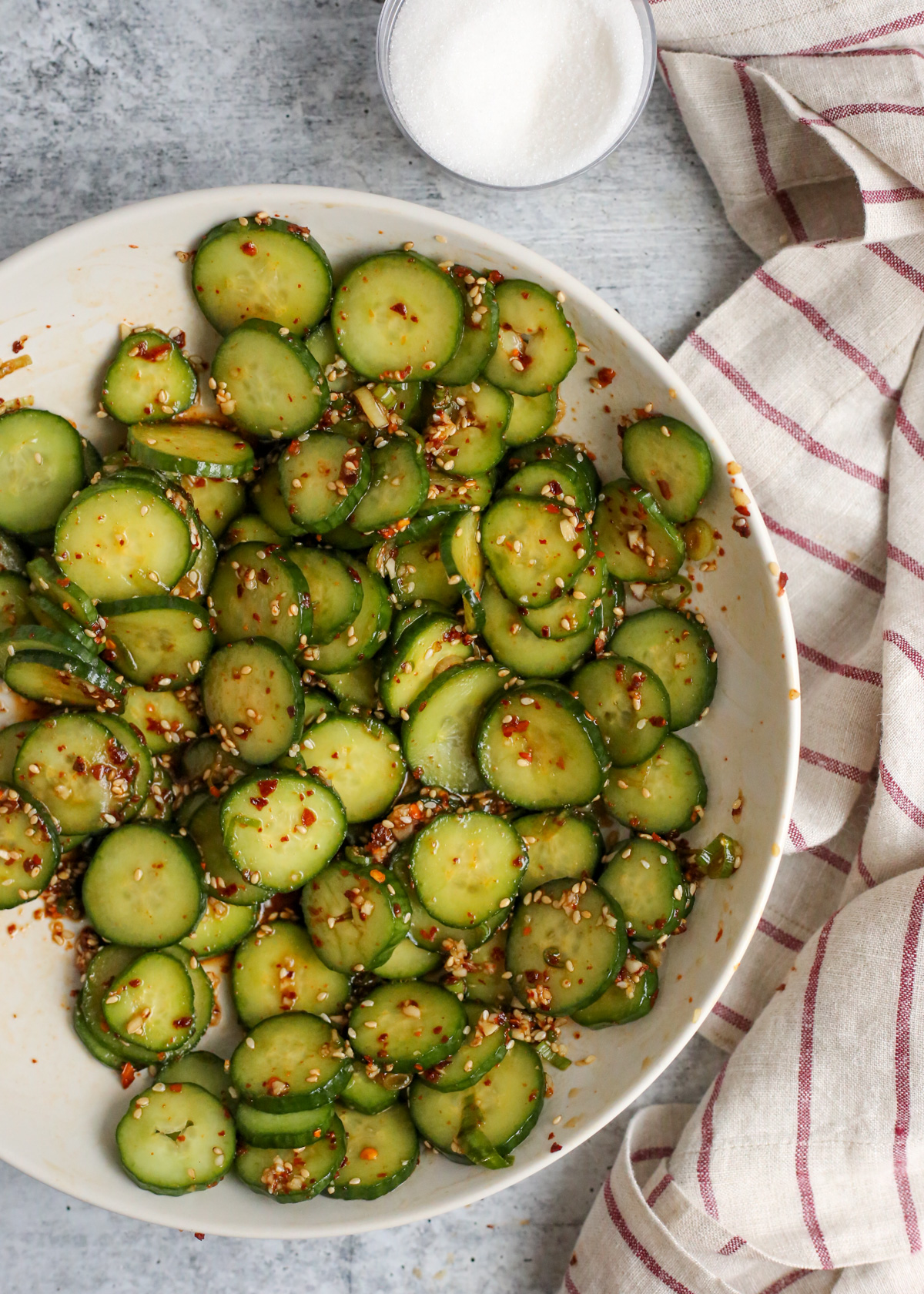 Overhead view of oi muchim, spicy korean cucumber salad, in a white serving dish with a striped linen and small jar of white sugar nearby