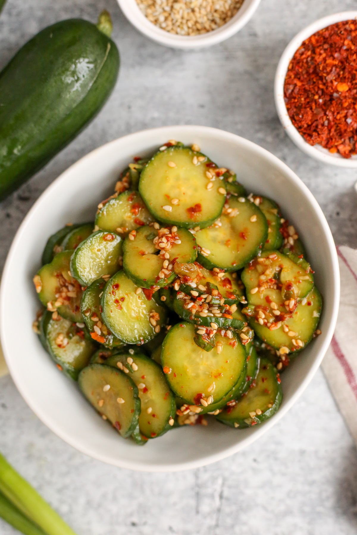 Overhead view of oi muchim, or spicy Korean cucumber salad, in a small white serving dish on a kitchen countertop, surrounded by a cucumber, ramekins of toasted sesame seeds and gochugaru, and a piece of green onion