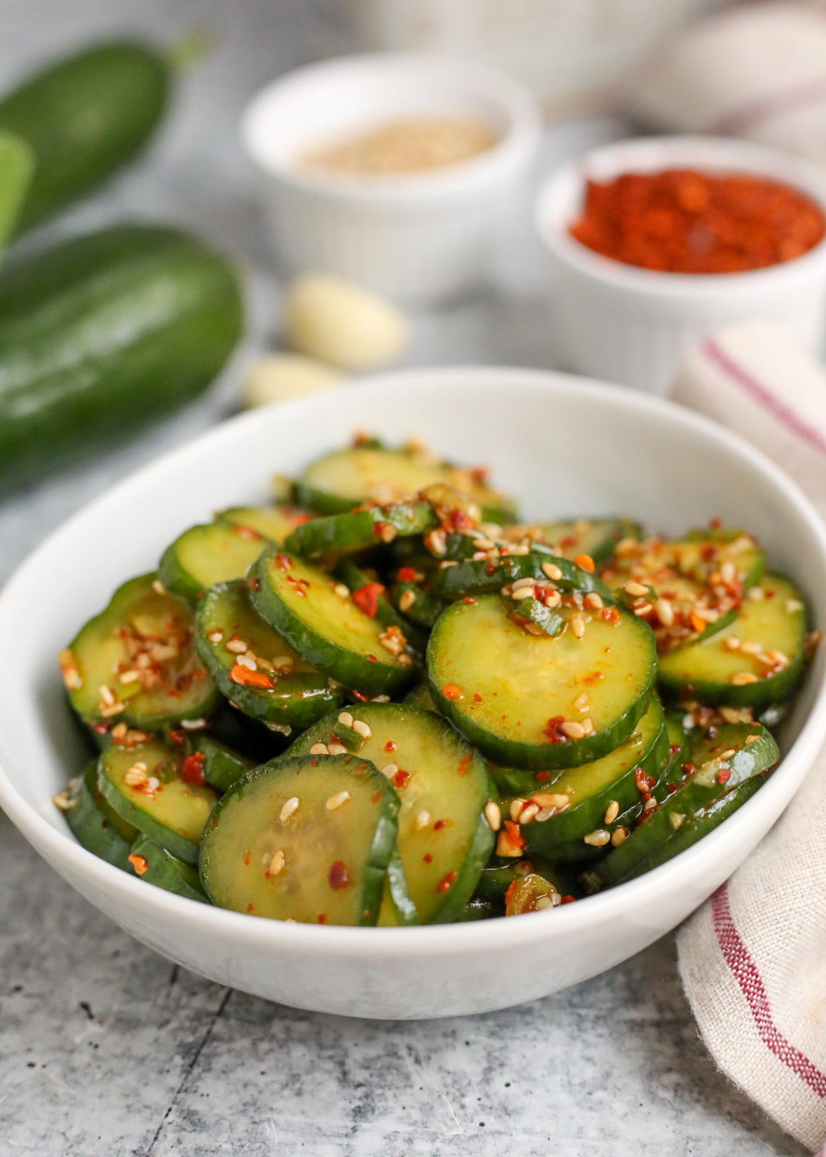 Side angle view of a small white ceramic dish filled with sliced cucumbers, seasoned and garnished with toasted sesame seeds and small red pepper flakes