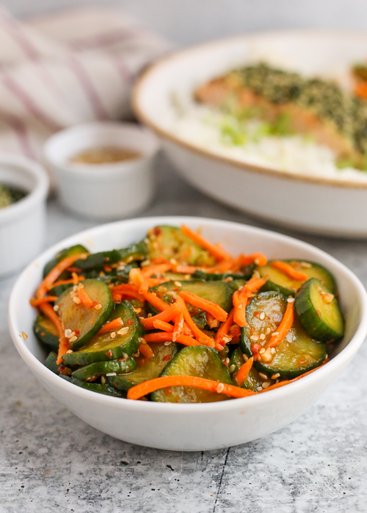 Side angle view of sliced cucumbers and matchstick carrots, seasoned and served in a small white dish with a main course displayed in a blurred background