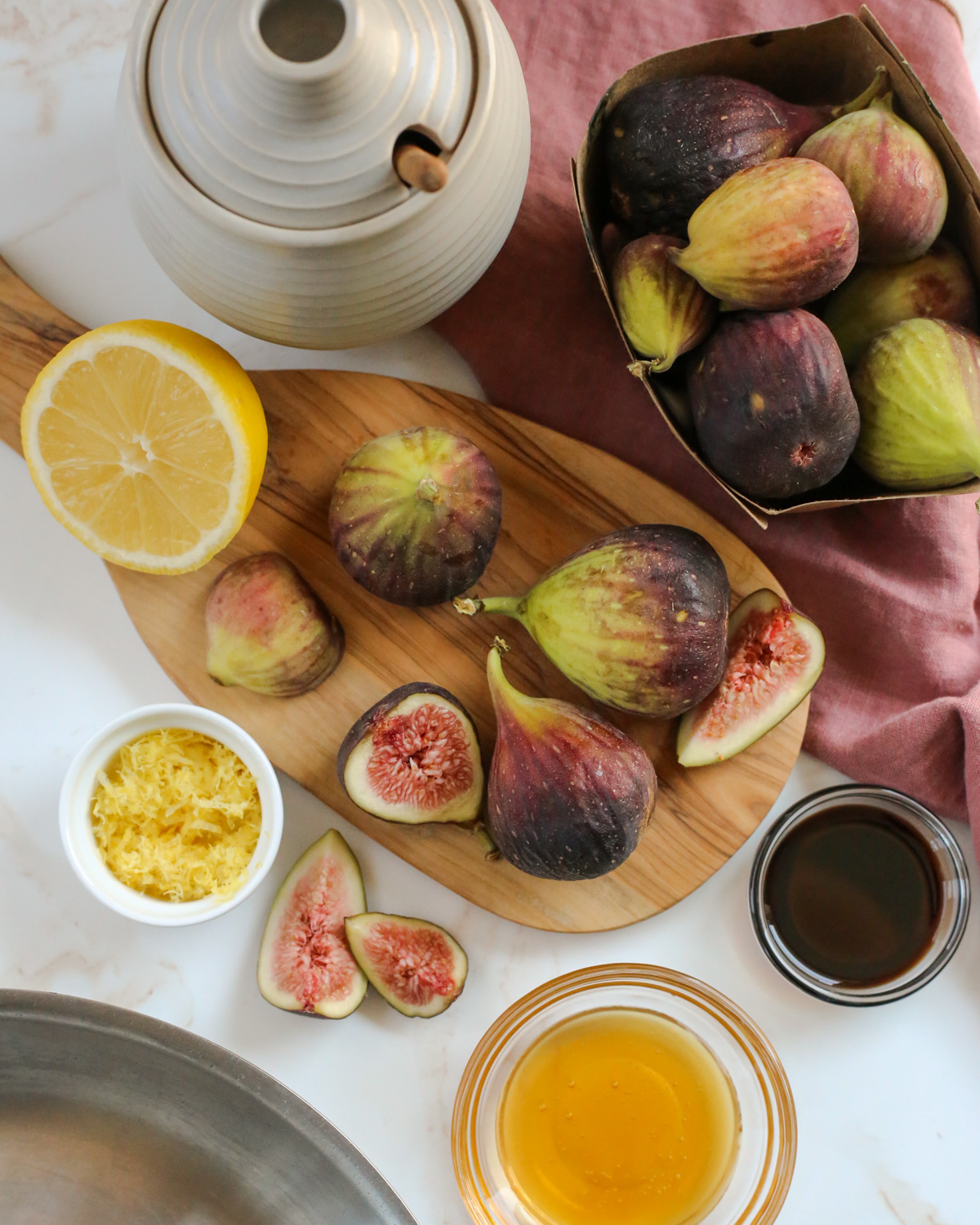 Overhead view of a kitchen scene, with fresh figs arranged on a wooden serving platter alongside small glass ramekins holding honey, balsamic vinegar, and lemon zest, with a stainless steel skillet partially in frame in the bottom left corner