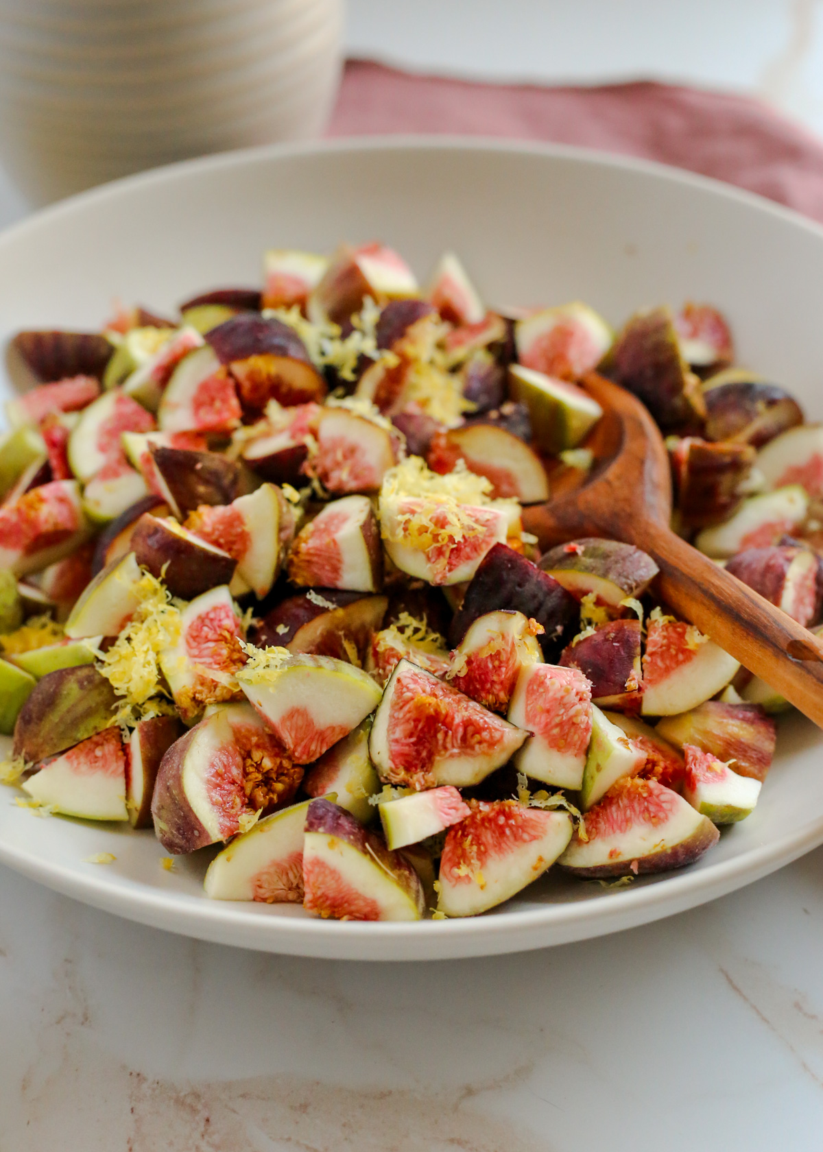 Angled view into a large shallow bowl holding fresh figs that have been sliced and chopped into small pieces. Lemon zest is visible on top and a wooden spoon rests on the side of the bowl
