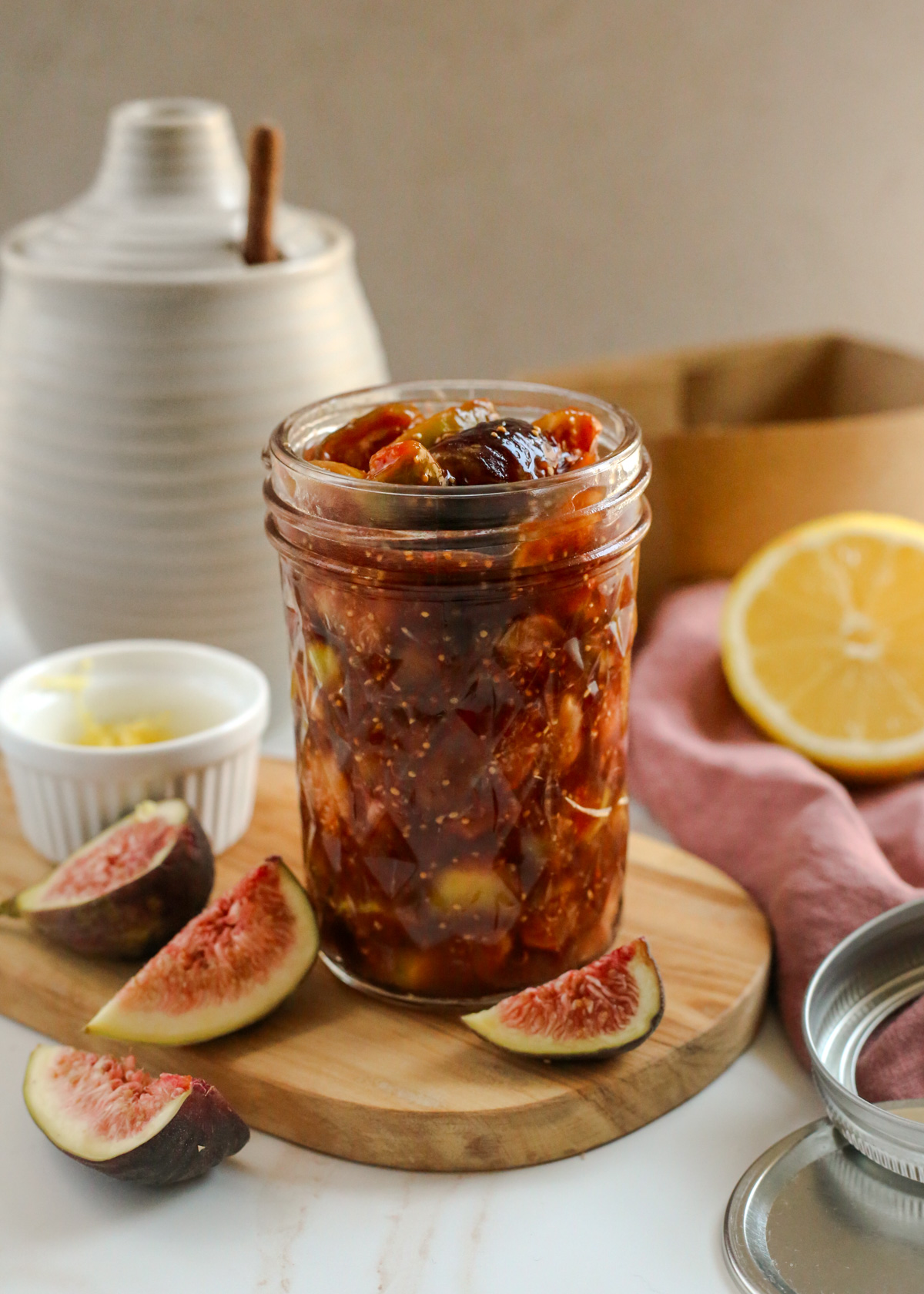 A close view of a balsamic fig compote recipe in a mason jar, with chunks of sweet balsamic glazed figs visible throughout, and additional ingredients and kitchen tools scattered in the background