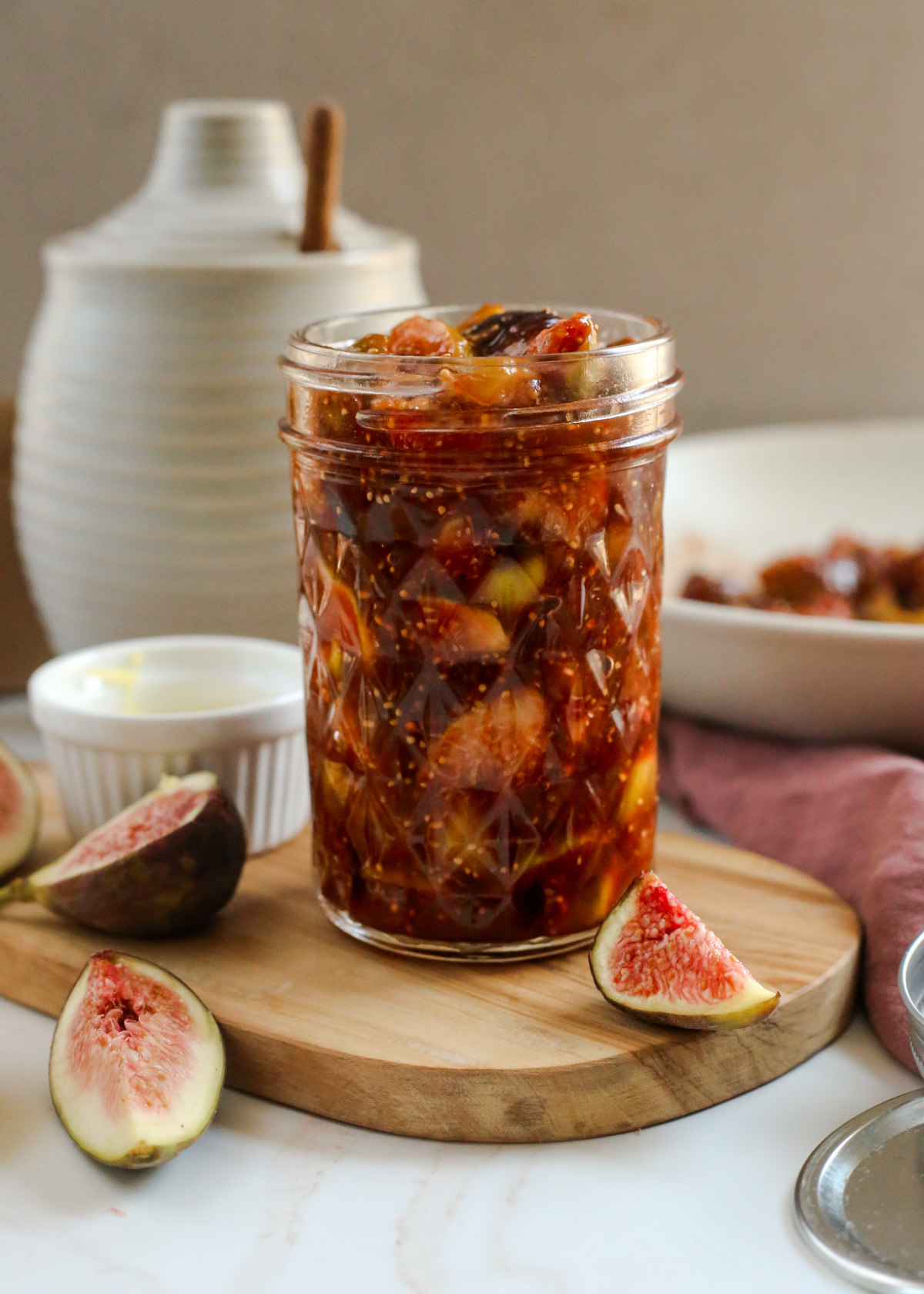 A freshly made balsamic fig compote is displayed in a mason jar on a wooden serving platter, with additional fresh figs scattered around it, softly lit from natural light from a kitchen window