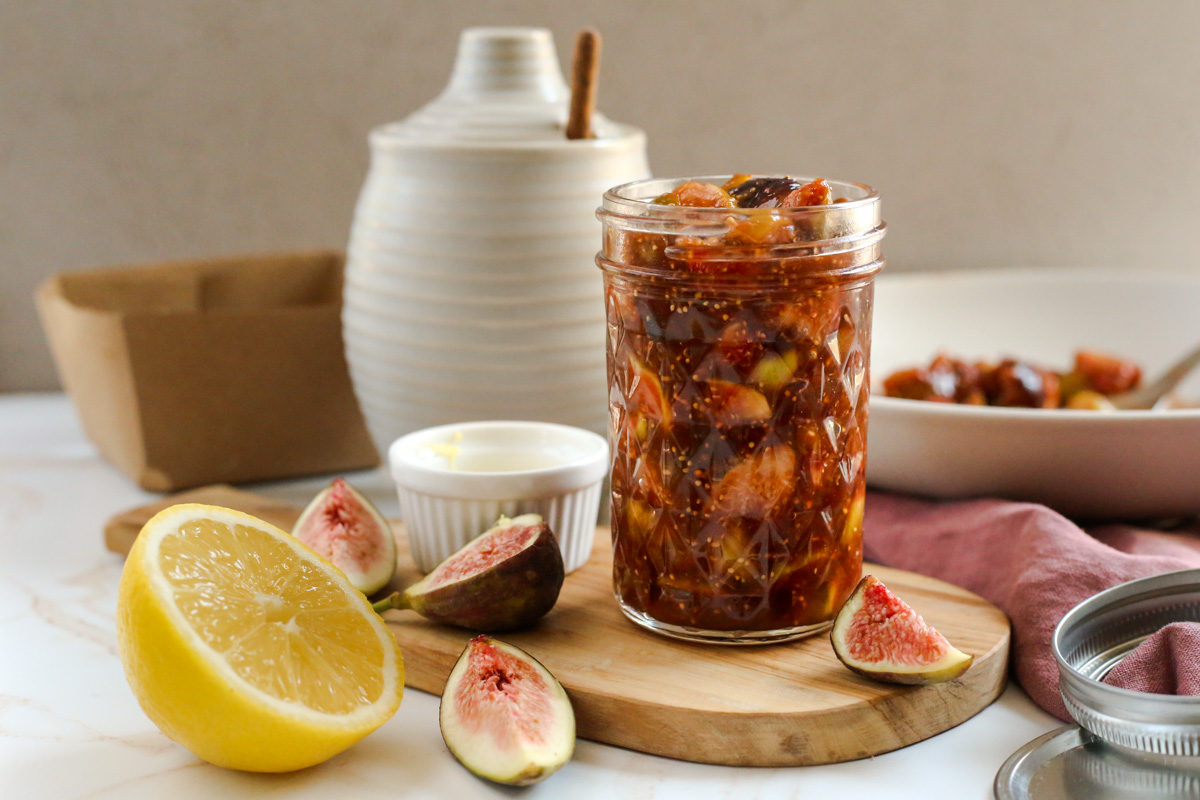 Lamdscape image of a fresh fig compote displayed in a small mason jar on a wooden serving platform, with a honey jar, lemon, and sliced figs arranged around it