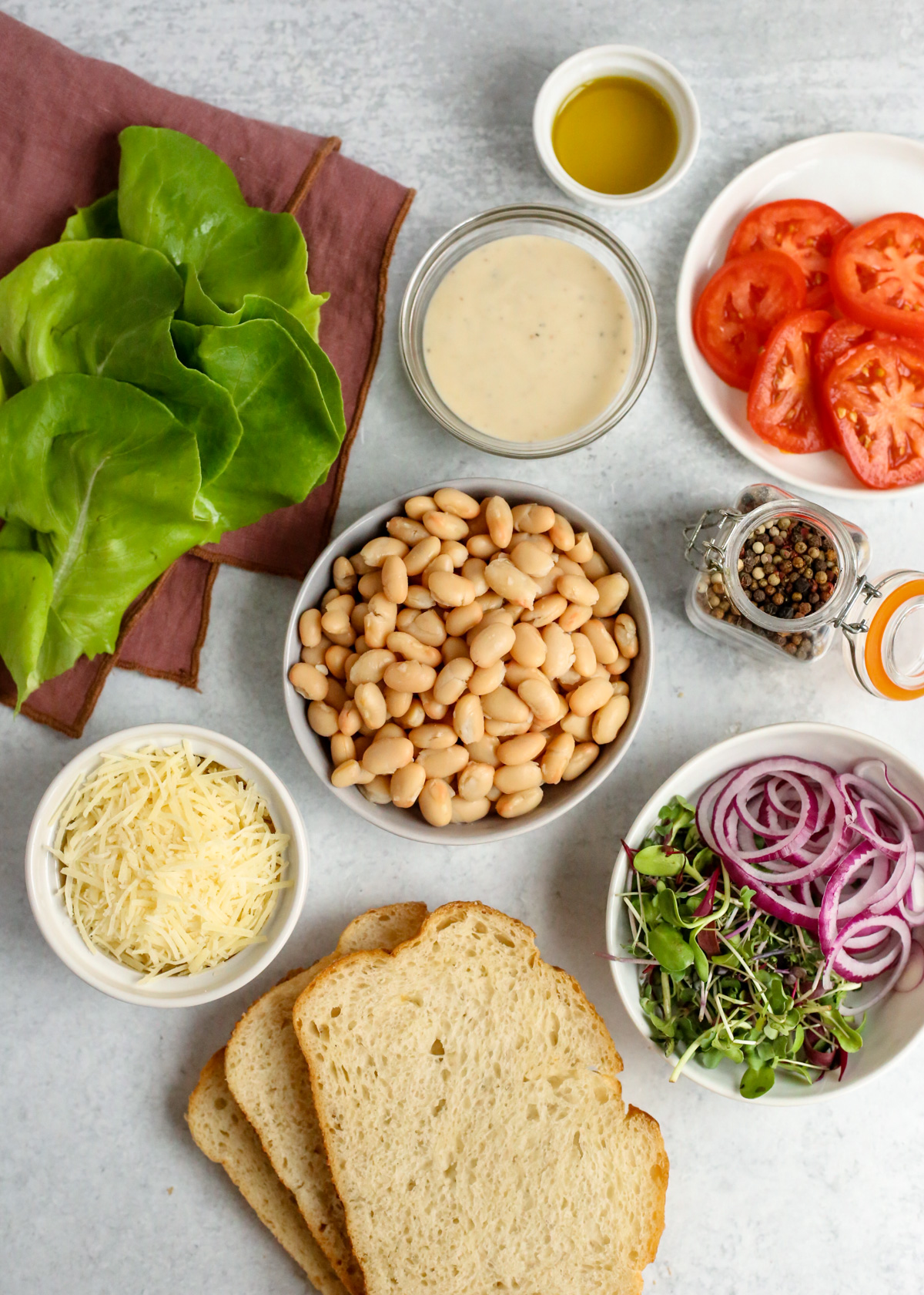 The ingredients needed to make caesar beans on toast arranged on a kitchen countertop in small dishes, including canned white beans, shredded parmesan cheese, sliced red onion and tomatoes, caesar dressing, lettuce, and slices of sourdough bread