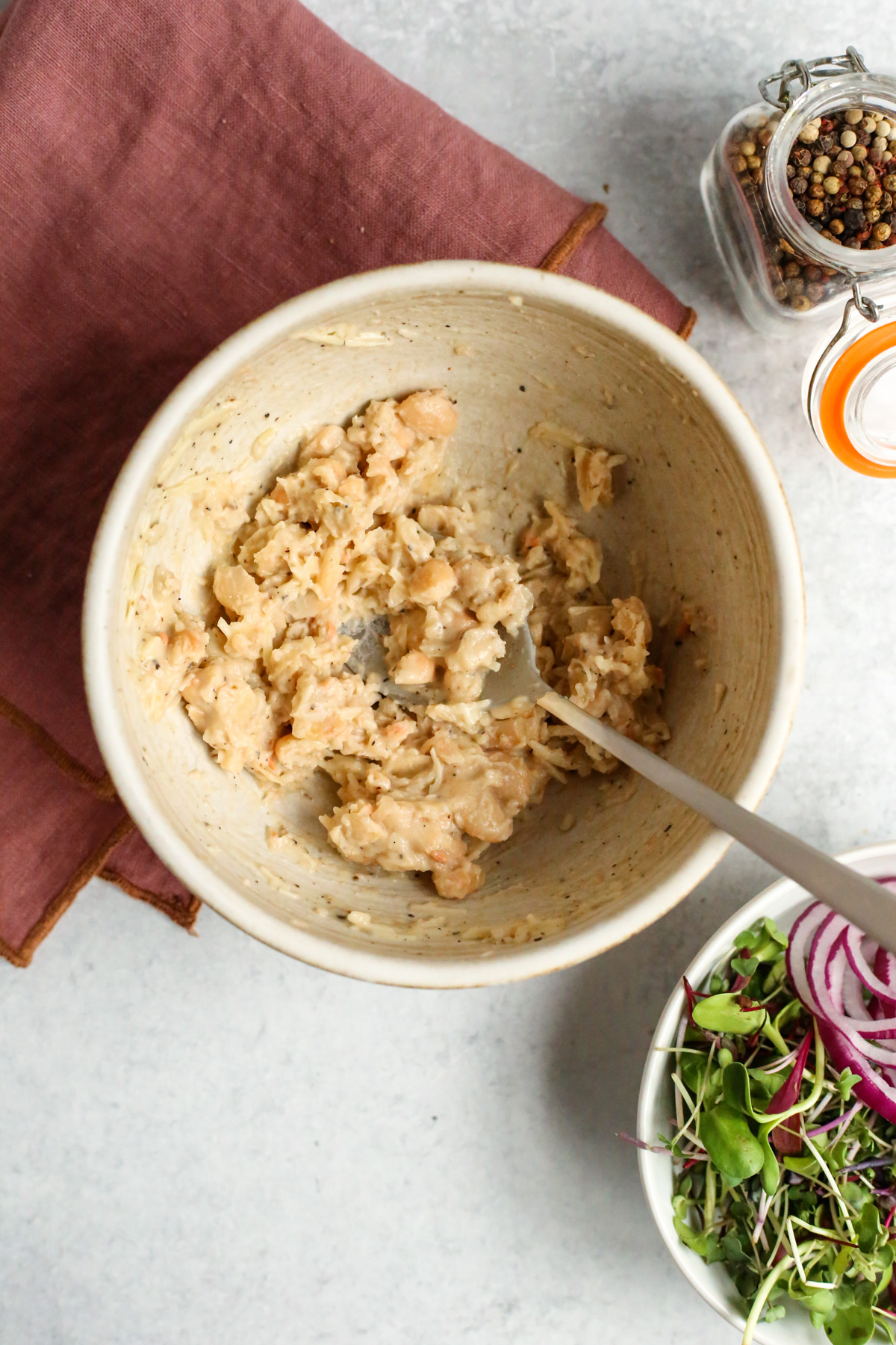 Overhead view of a handmade ceramic bowl containing smashed white beans, mixed with caesar salad dressing, black pepper, and shredded parmesan cheese. A silver spoon rests in the bowl, and the smashed beans have a chunky, creamy consistency