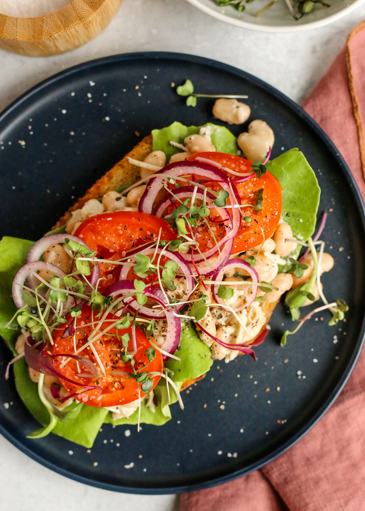 Overhead view of Caesar Beans on Toast, with sourdough bread topped with white beans coated in caesar dressing, stacked with fresh lettuce, sliced tomatoes and red onions, micro greens, and black pepper for garnish
