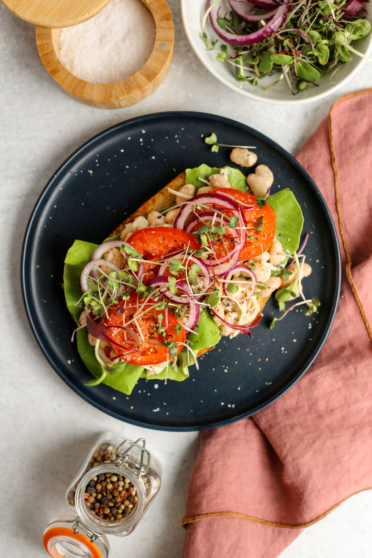 Overhead view of a Caesar Beans on Toast recipe, with one slice of toasted bread topped with lettuce, caesar beans, sliced tomatoes, red onions, and garnish on a navy blue ceramic plate on a kitchen countertop