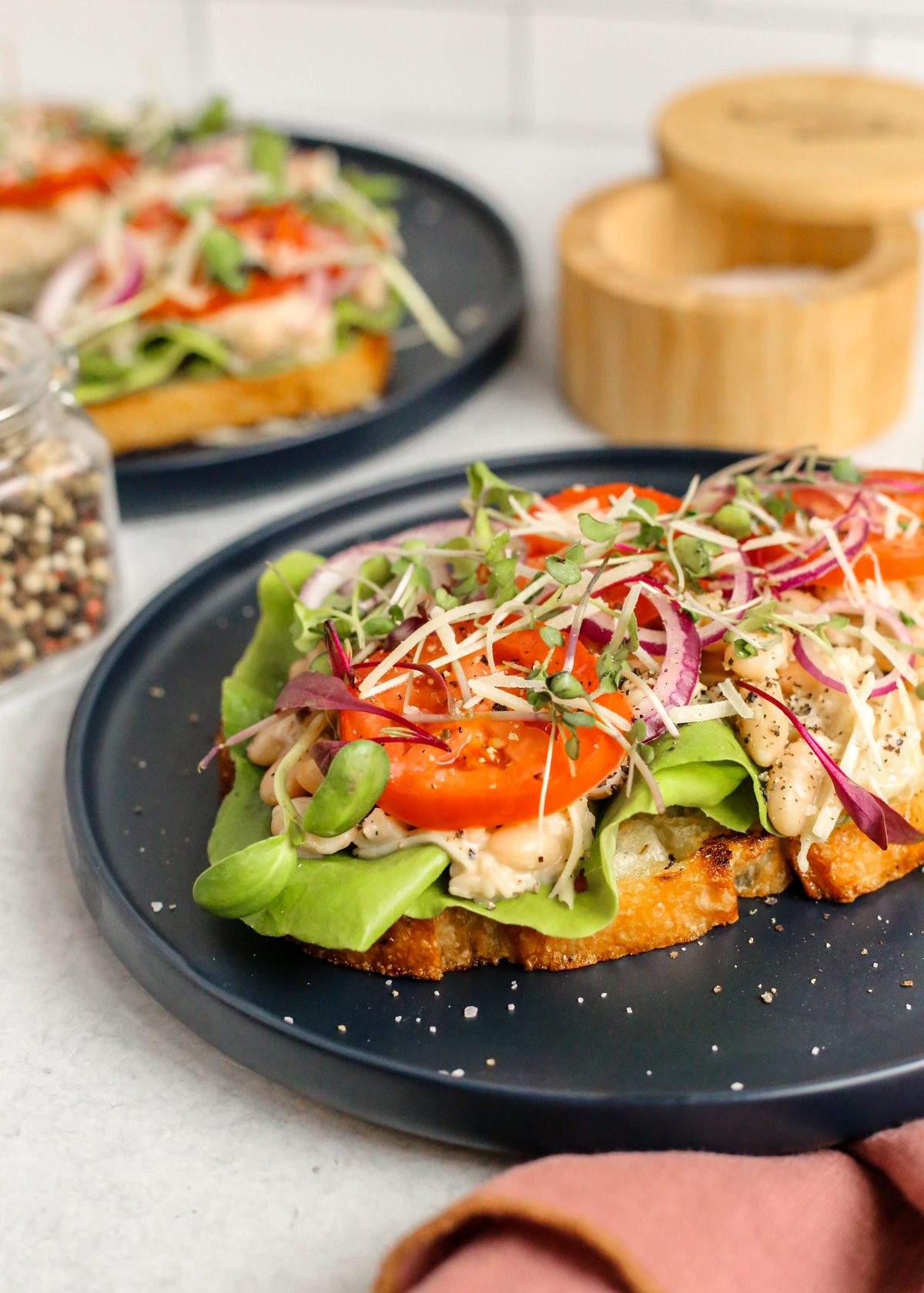 Styled image of smashed white beans coated in caesar dressing on open-faced toast with lettuce, tomatoes, and red onion, garnished with black pepper, salt, and micro greens, served on a navy blue ceramic dish on a kitchen countertop