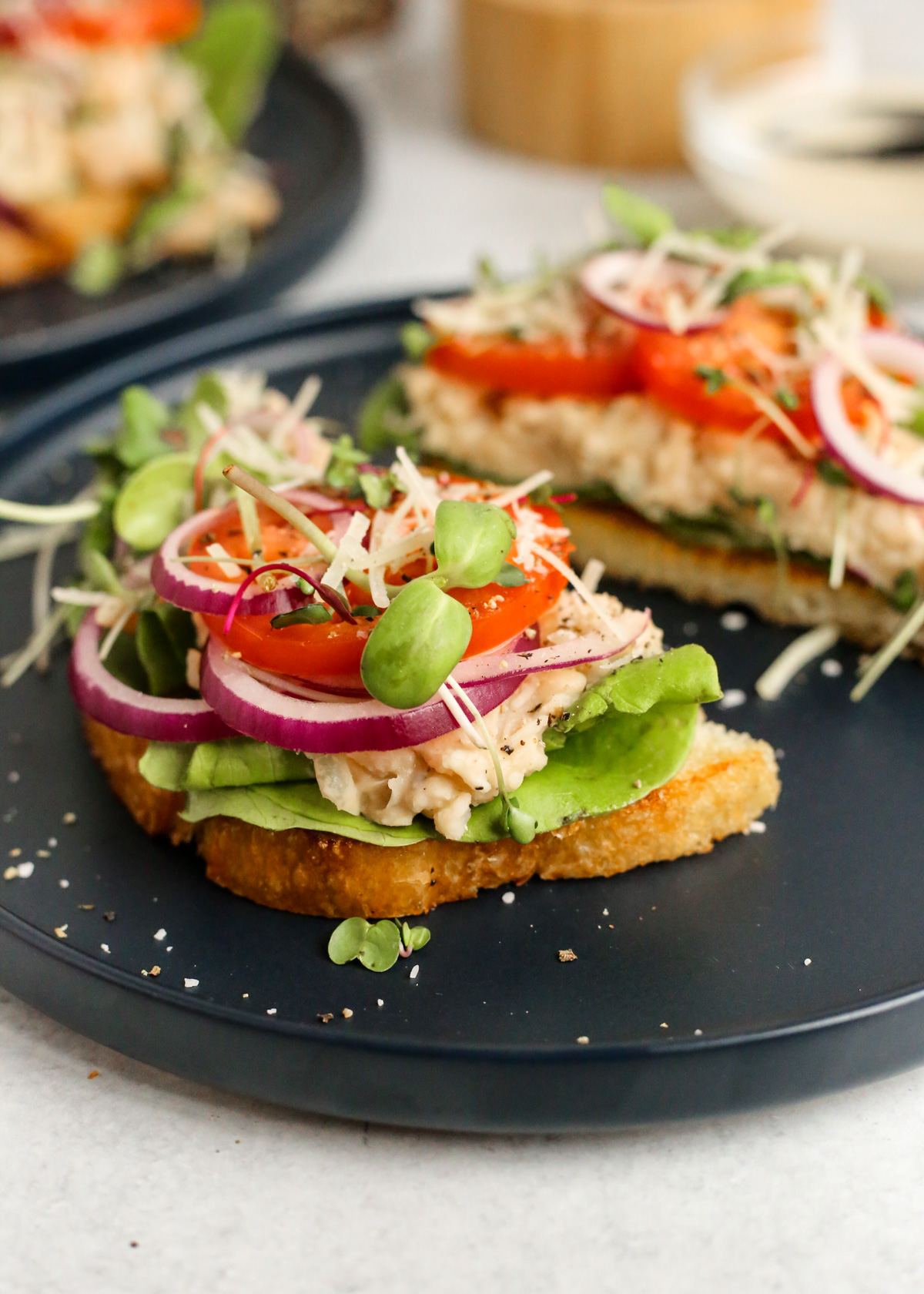 A thick slice of toasted sourdough is topped with fresh lettuce, caesar beans, sliced tomatoes and red onion, micro greens, and shredded parmesan cheese, with the toast sliced in half to showcase the layered ingredients