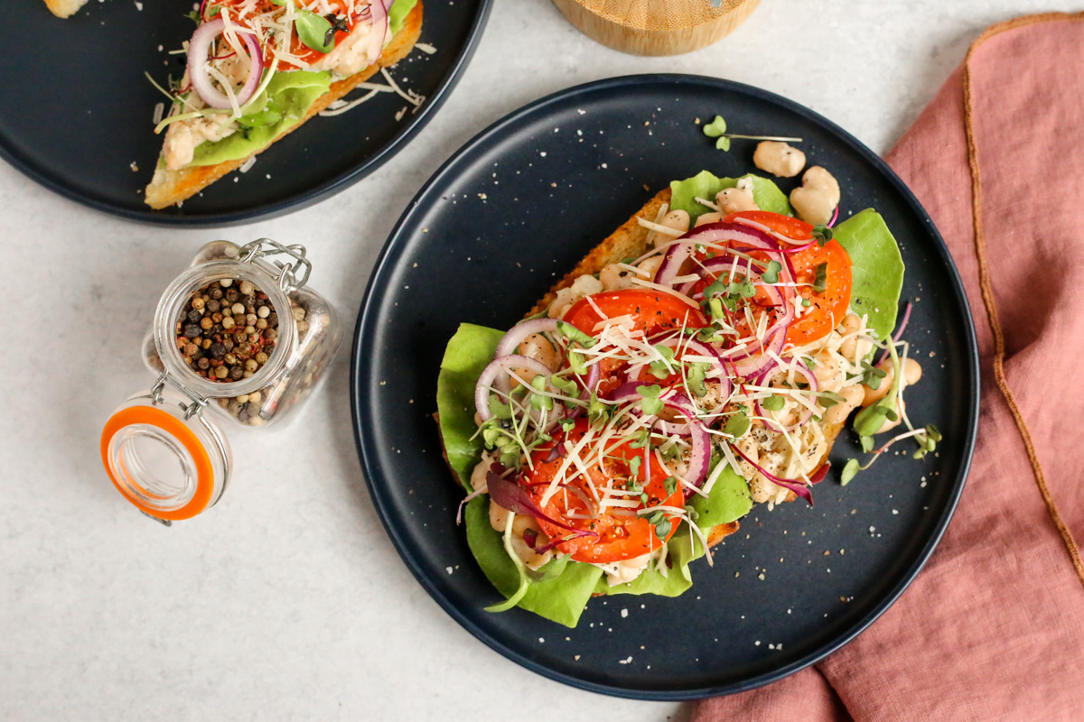 Landscape oriented photo of styled caesar beans on toast, served on a navy blue ceramic dish on a kitchen countertop. The toast is layered with white beans coated in caesar dressing, fresh lettuce, sliced tomatoes and red onion, micro greens, and black pepper for garnish