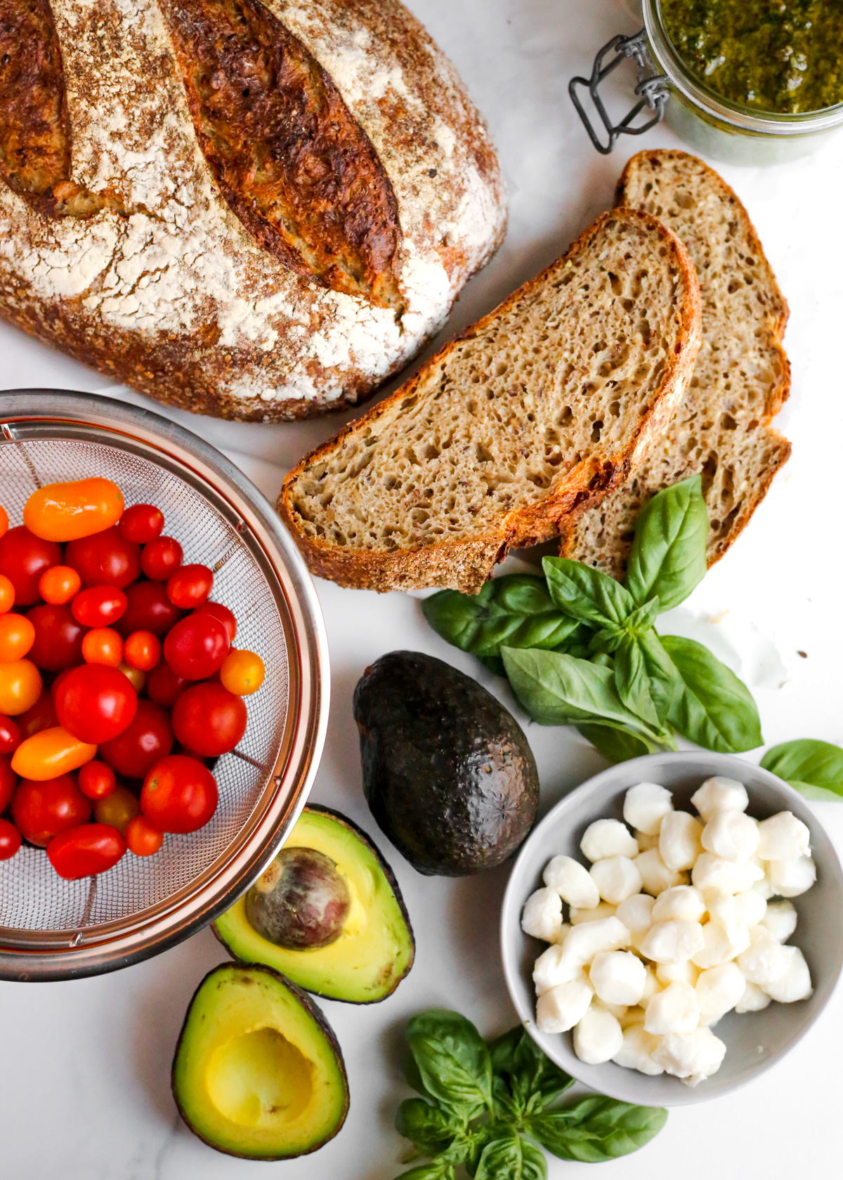 Overhead view of the ingredients needed to make cparese avocado toast, including thick slices of multigrain bread, pesto sauce in a glass jar, cherry tomatoes, a ripe avocado, mozzarella cheese, and fresh basil