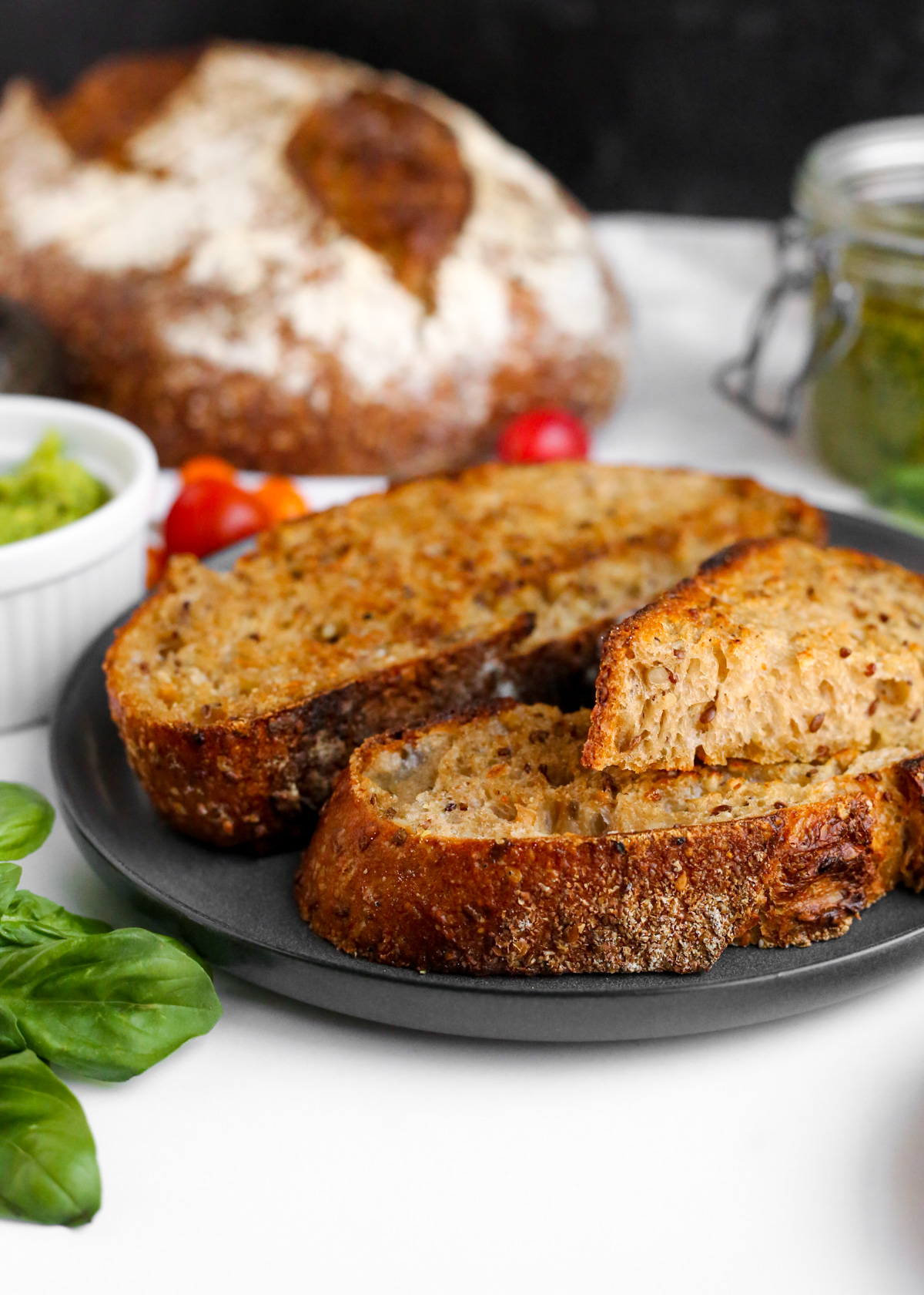 A closeup view of toasted slices of multigrain bread, appearing golden brown and crispy on the outside. Cherry tomatoes, fresh basil, mashed avocado, and a jar of pesto sauce are included in the background