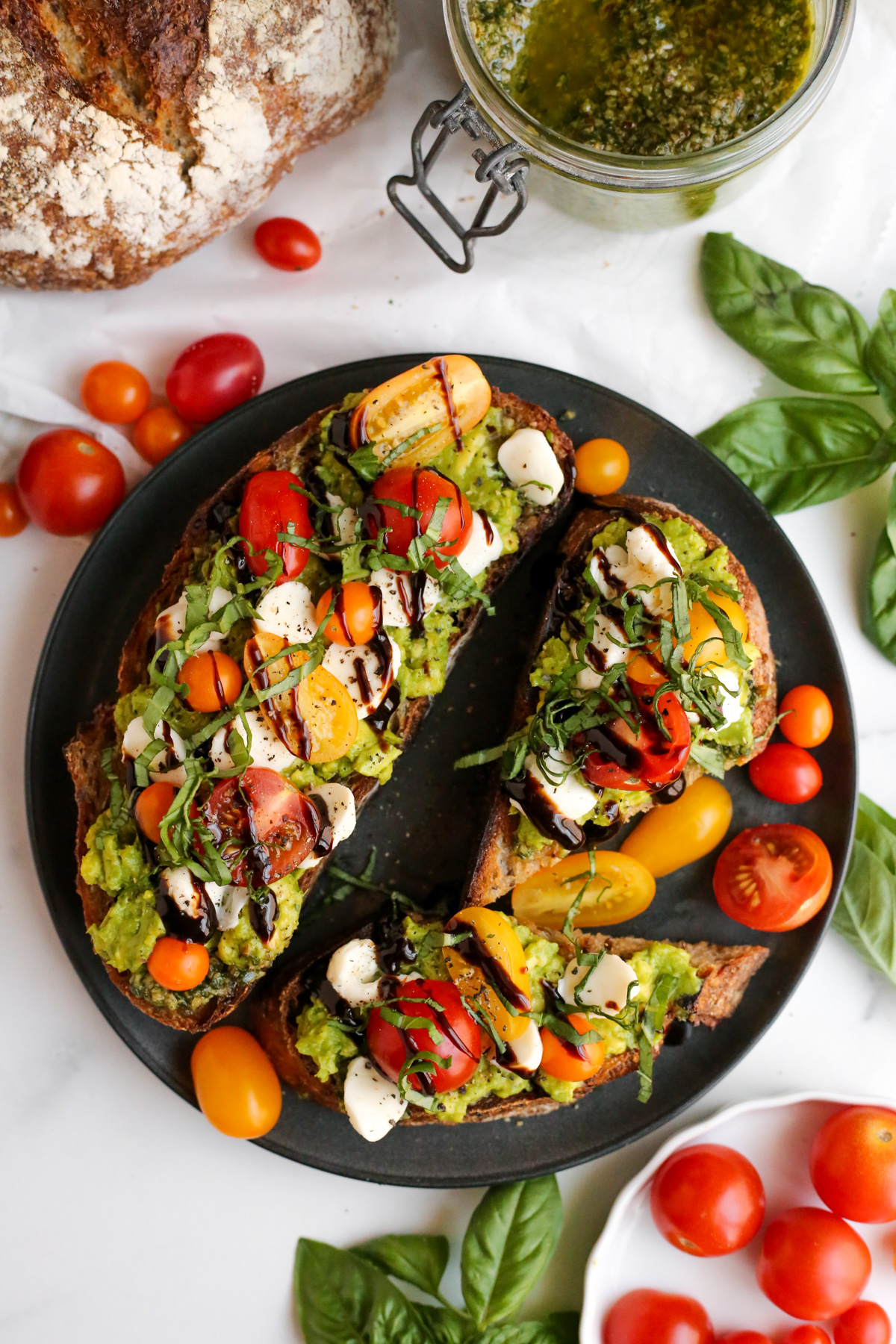 Overhead view of two thick slices of toasted multigrain bread, smeared with pesto sauce and stacked with a layer of mashed avocado, sliced cherry tomatoes and mozzarella cheese, fresh basil, and drizzled with balsamic glaze. The avocado toast is served on a black ceramic dish on a kitchen countertop