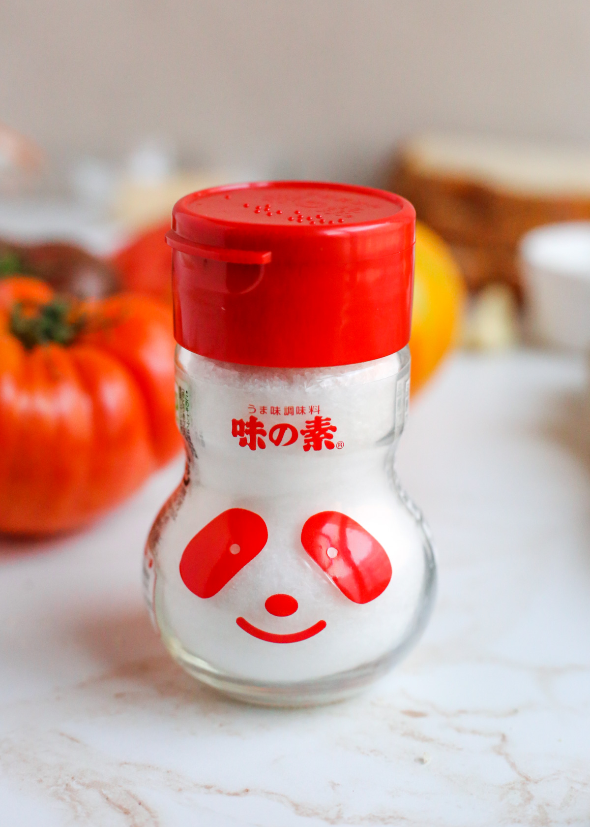 A panda-shaped shaker bottle with a red lid and lettering containing MSG is displayed on a kitchen countertop with fresh heirloom tomatoes in the background