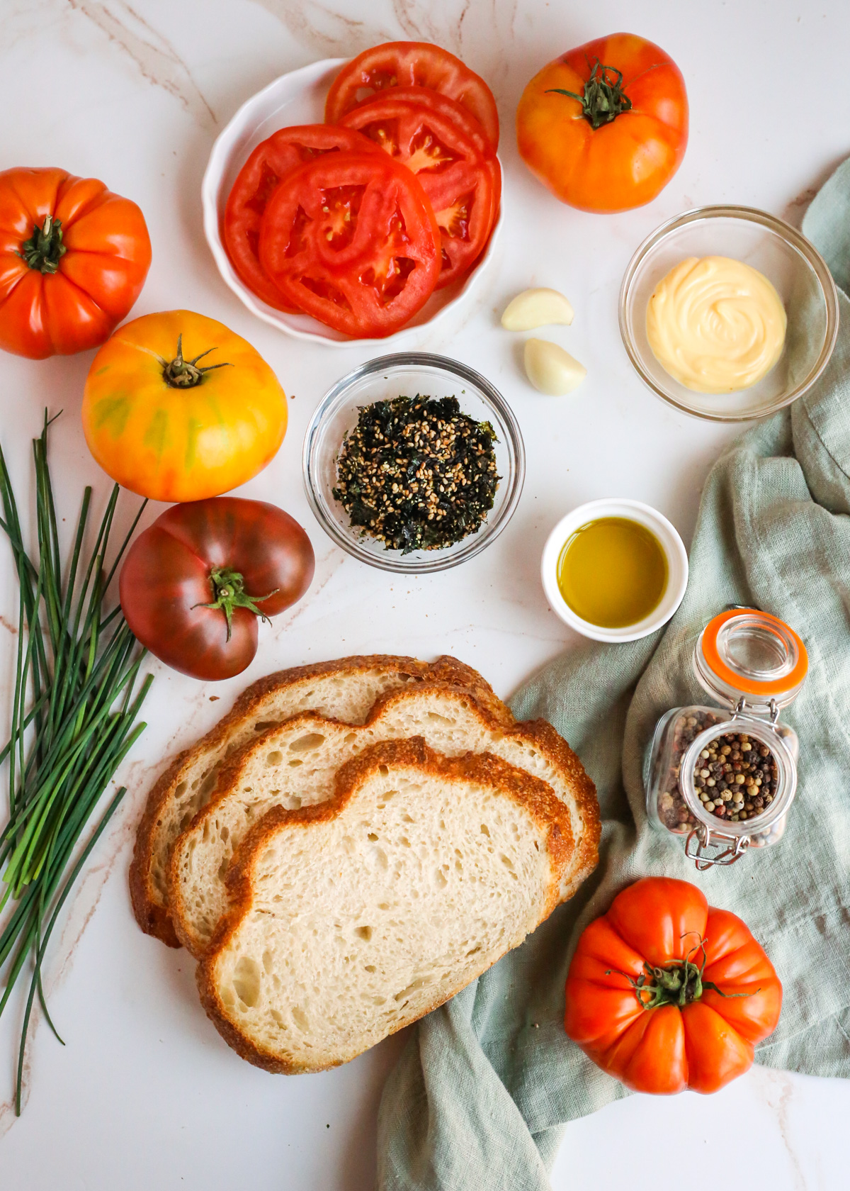 Flatlay style image of all the ingredients needed to make a Furikake Tomato toast recipe, displayed in various small prep bowls or ramekins on a beige countertop