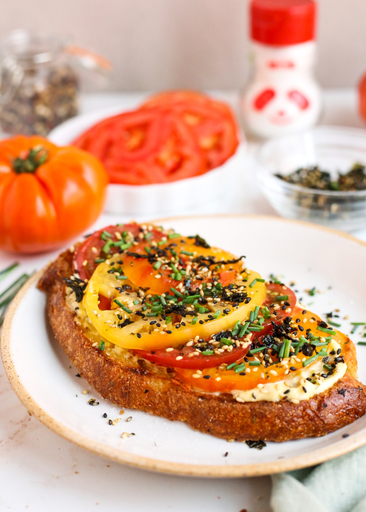 A breakfast scene with a dish containing a slice of Furikake Tomato Toast, displayed in front of heirloom tomatoes, a small dish of furikake, and a panda-shaped shaker bottle containing MSG