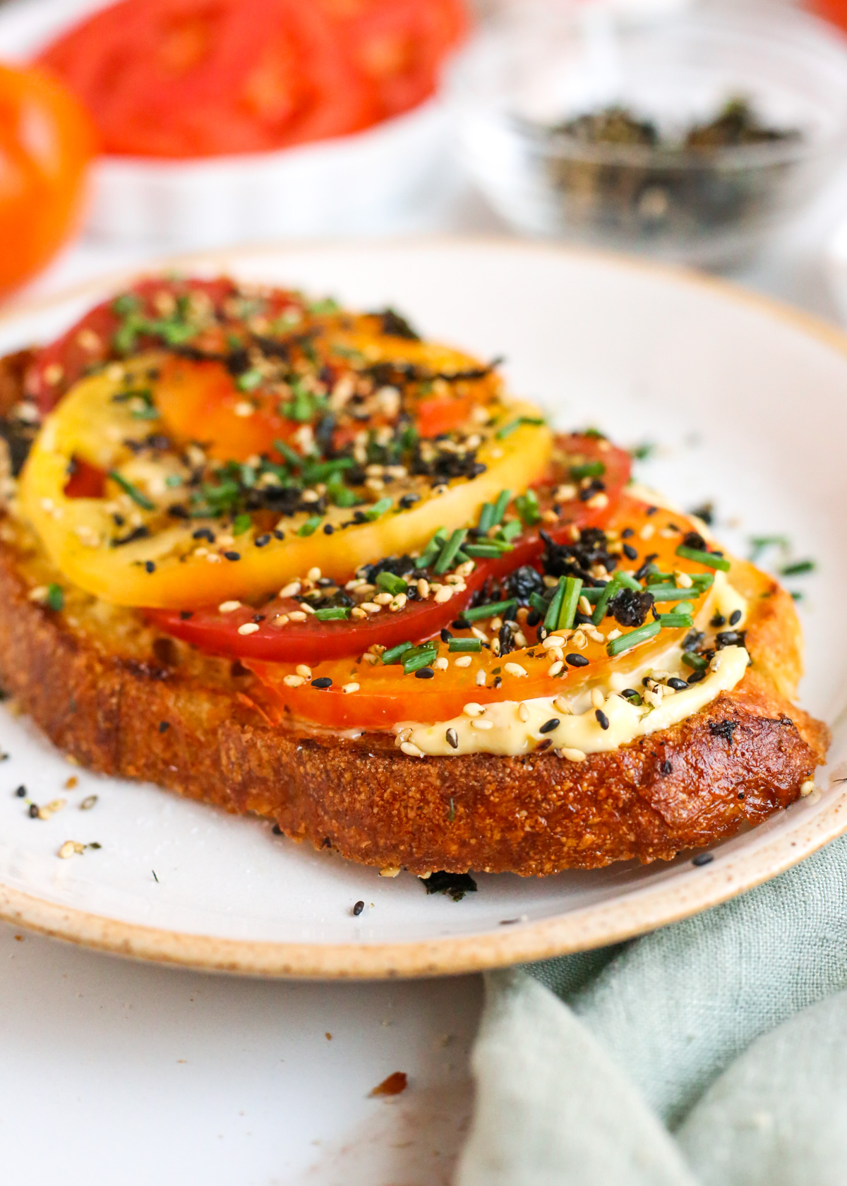 A side view of a slice of Furikake Tomato Toast, showing the layers of golden toasted sourdough bread, thick and creamy garlic mayo, thick slices of heirloom tomatoes, and a rustic garnish of furikake and chopped chives