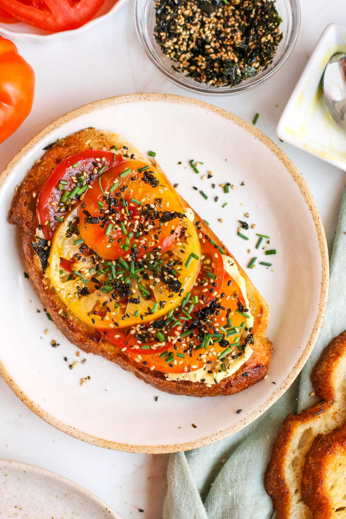 Overhead view of a slice of tomato toast on sourdough bread, with colorful red, orange, and yellow heirloom tomato slices, topped with furikake and chopped chives, served on a ceramic dish on a kitchen countertop