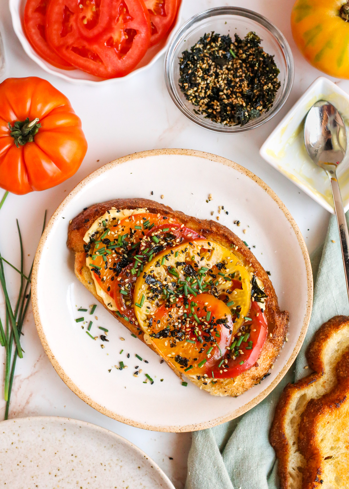 Overhead view of a plate with a slice of tomato toast, garnished with furikake and chopped chives, with additional tomatoes, chives, sourdough bread slices, and garlic mayo arranged nearby as it it was just made moments ago