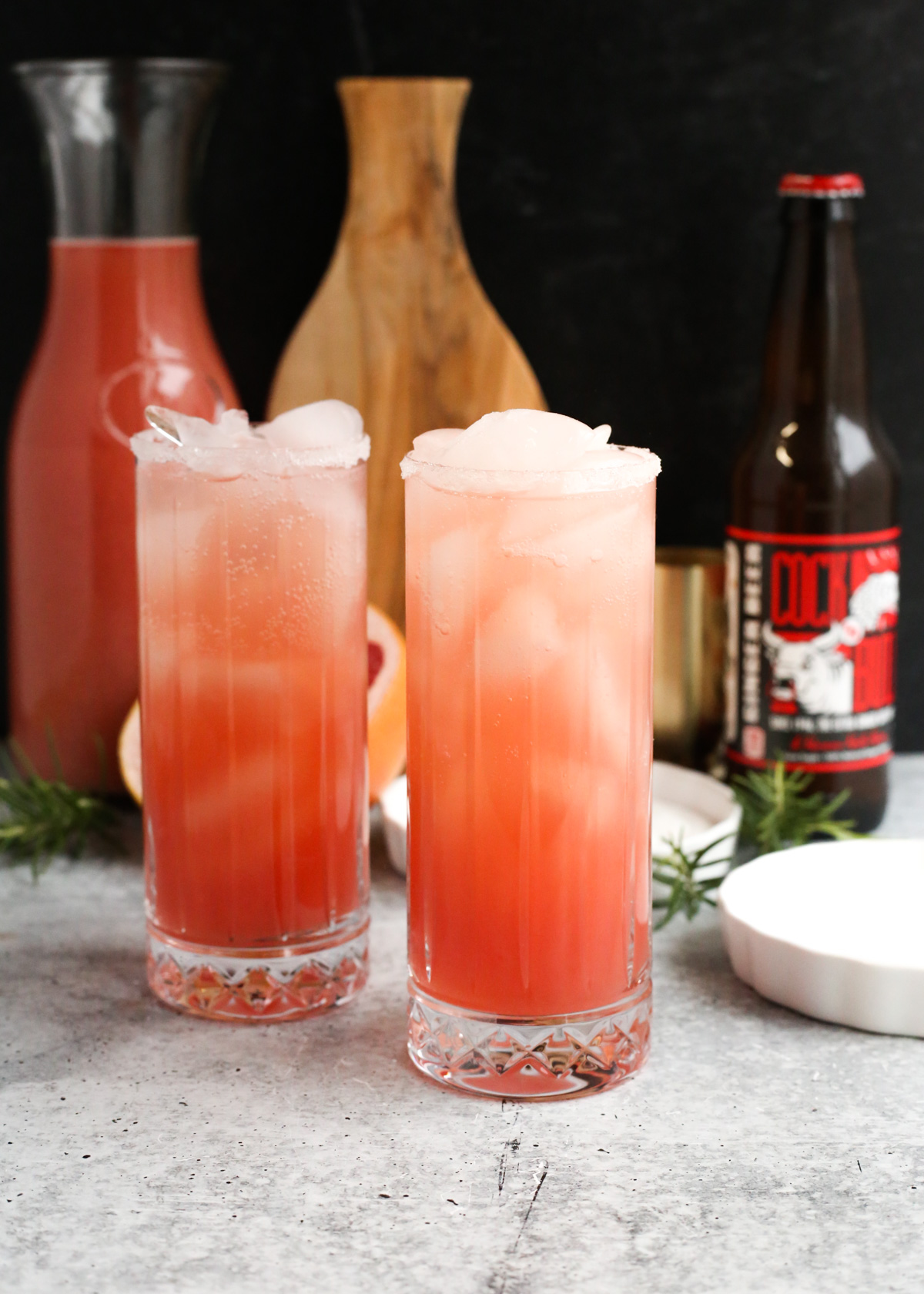 Two ginger beer paloma cocktails are served on a kitchen countertop, filled to the top of tall glasses with crushed ice and the garnishes visible in the background