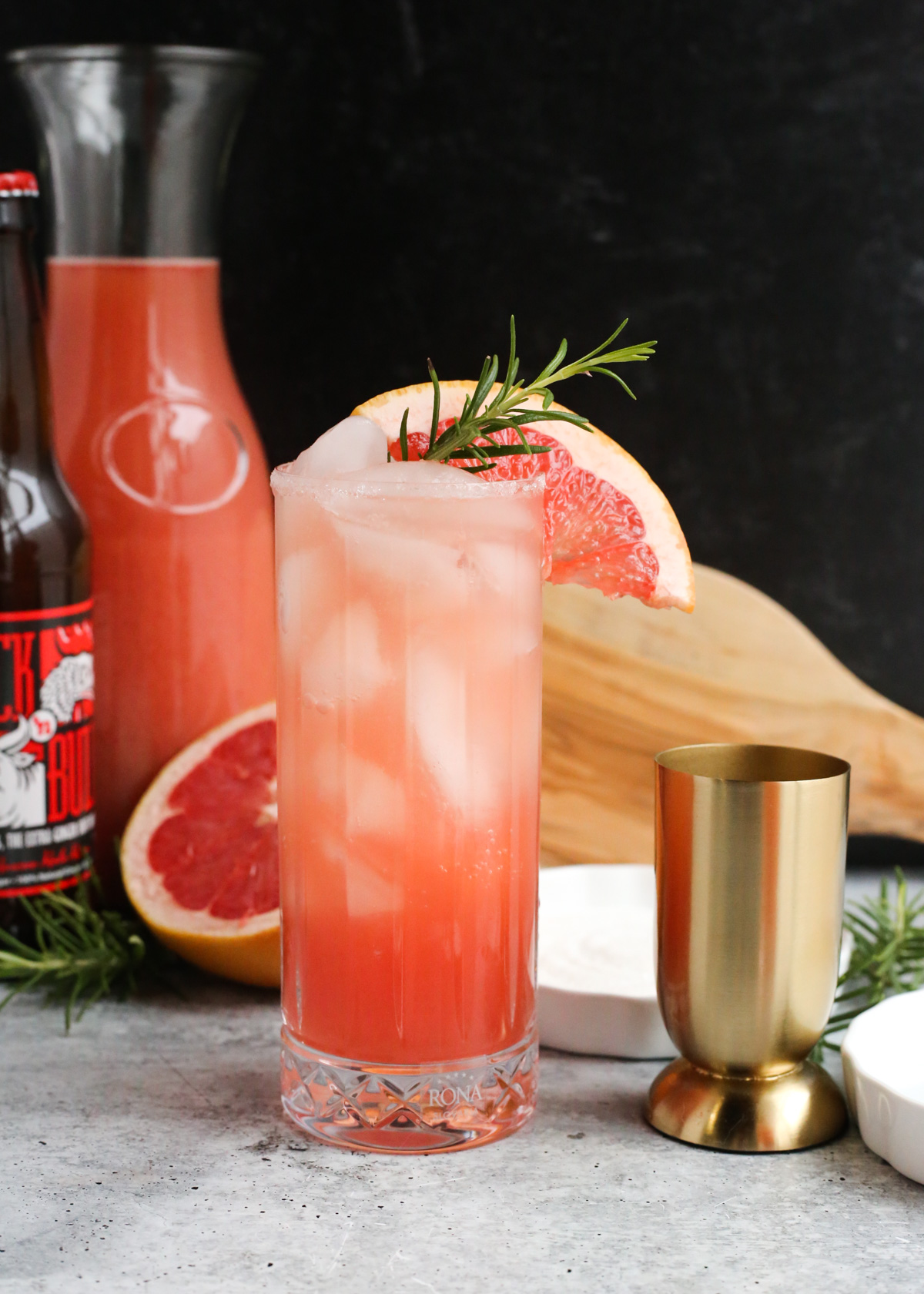 A ginger beer paloma cocktail is served in a highball glass and garnished with a sprig of rosemary and slice of grapefruit, with a bottle of ginger beer and carafe of fresh grapefruit juice in the background