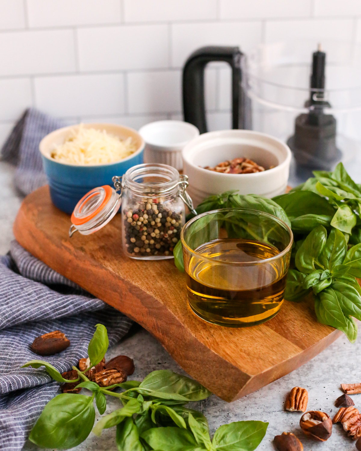 A wooden serving tray displays the ingredients needed for a homemade pesto without pine nuts, including olive oil, black pepper, parmesan cheese, salt and MSG, pecans, and fresh basil. The bowl of a food processor is visible in the background